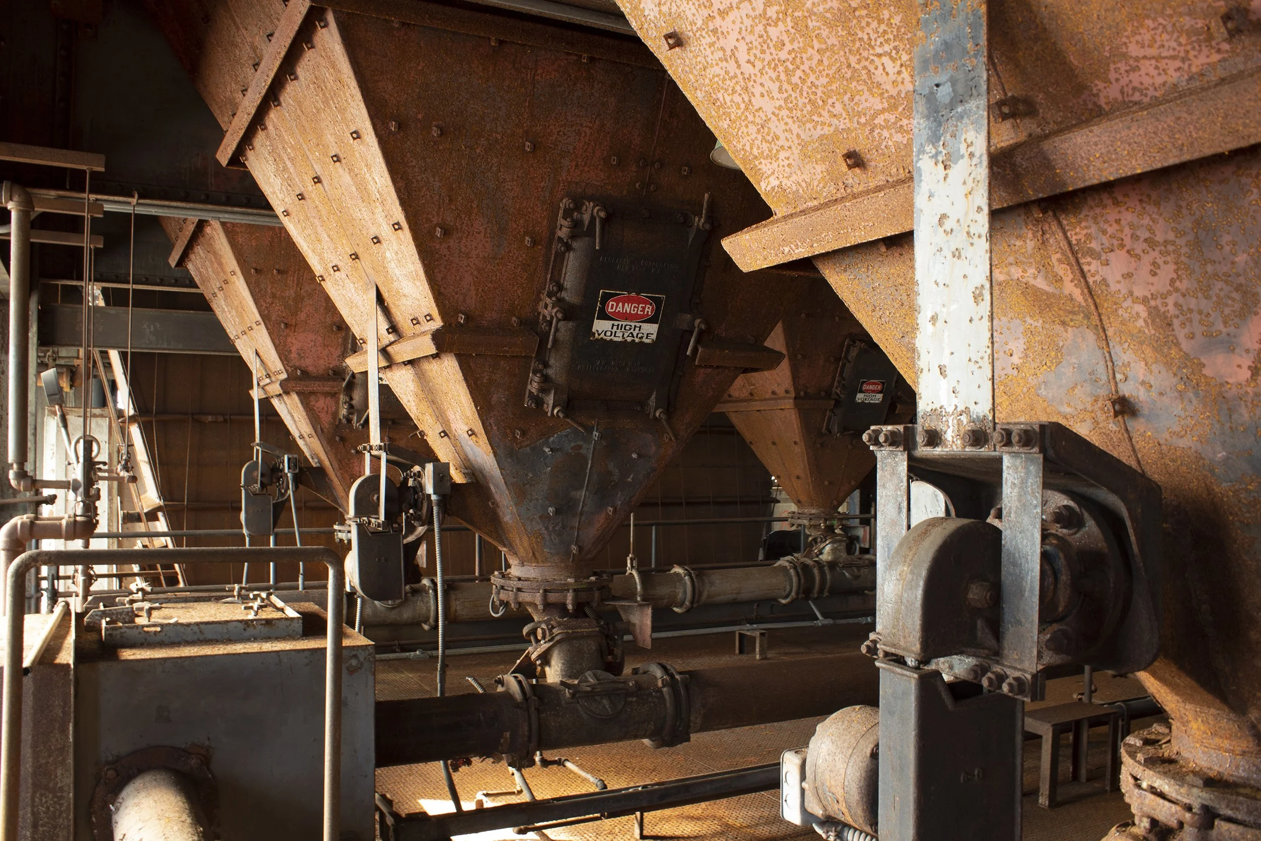 Rusty industrial machinery with pipes, valves, and a high voltage warning sign in an industrial setting.