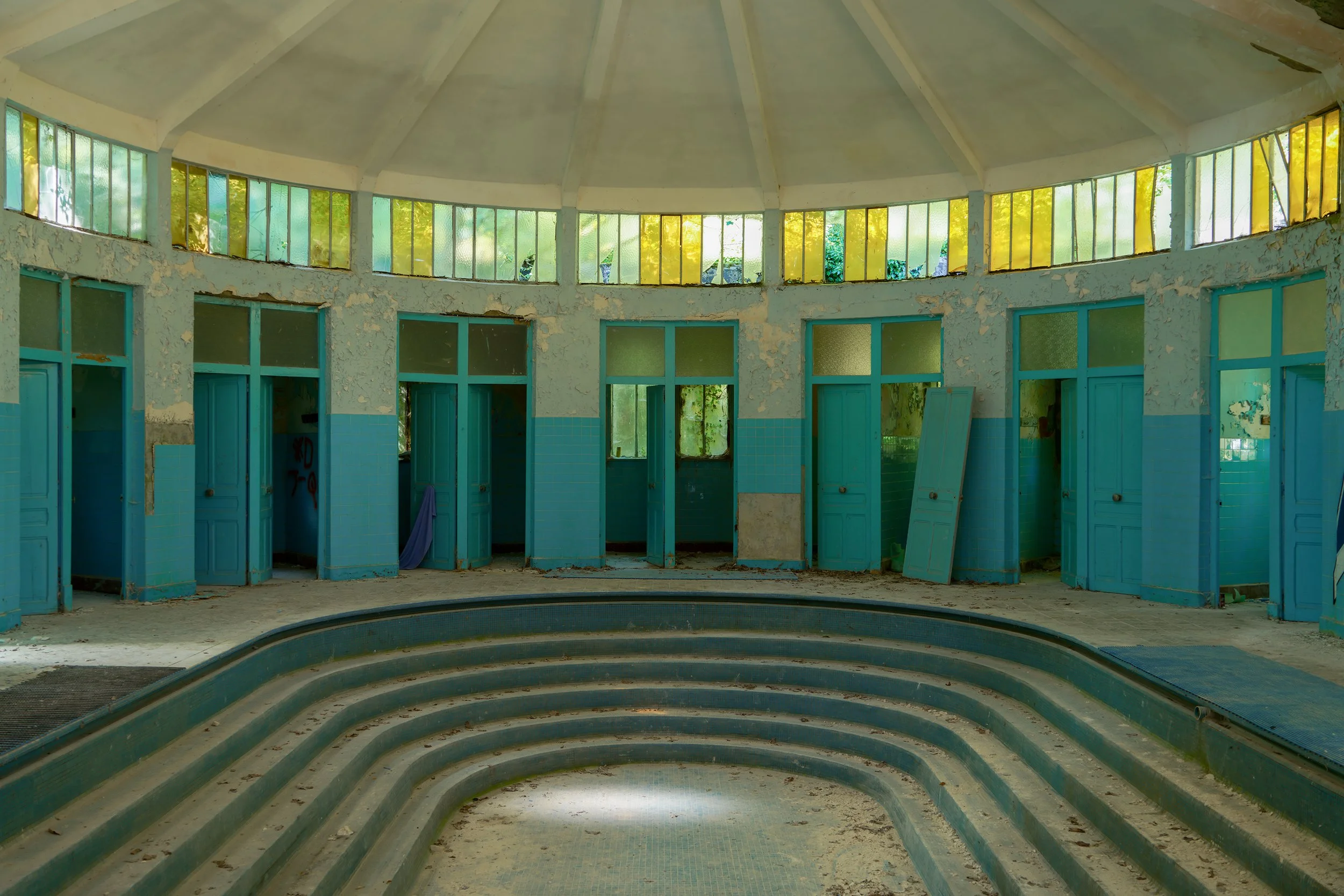 An abandoned public swimming pool with faded blue doors and peeling paint, surrounded by overgrown vegetation visible through dirty windows.