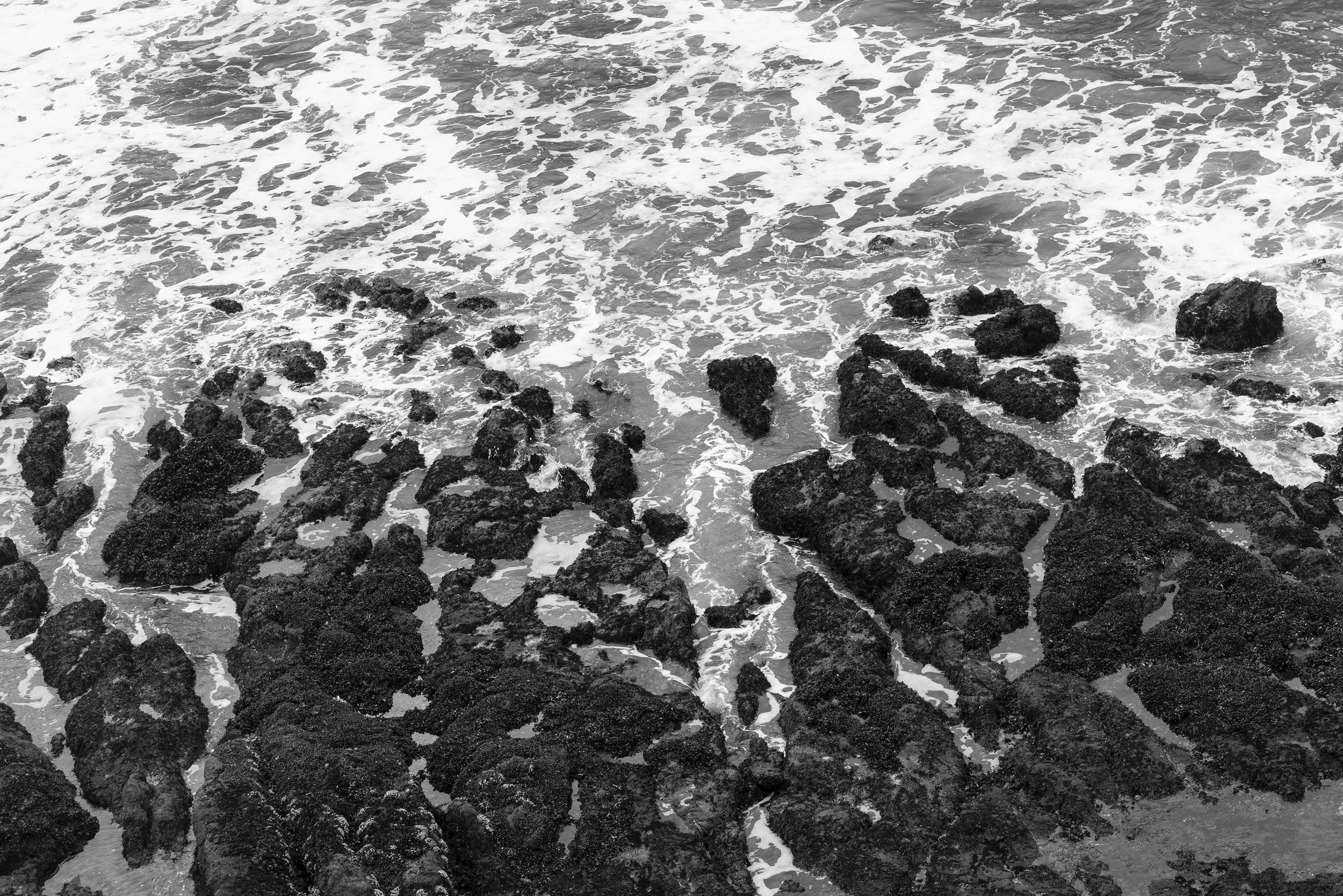 Black and white photograph of ocean waves hitting rocks along the shoreline.