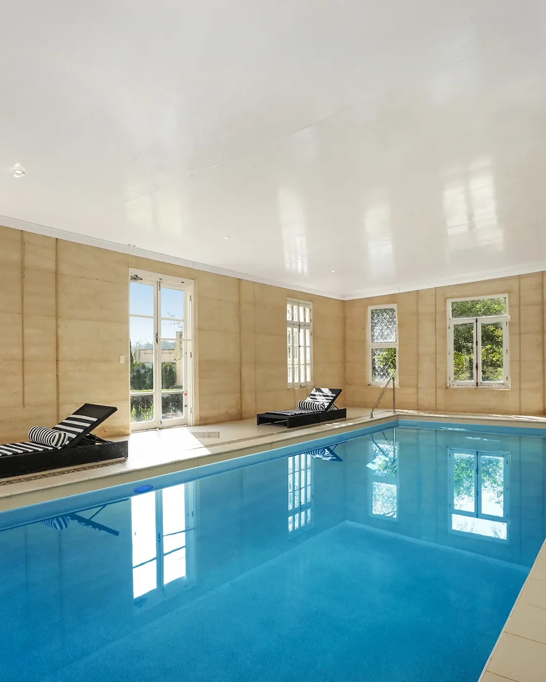 Indoor swimming pool with blue water, black and white striped lounge chairs, beige walls, and large windows letting in natural light.