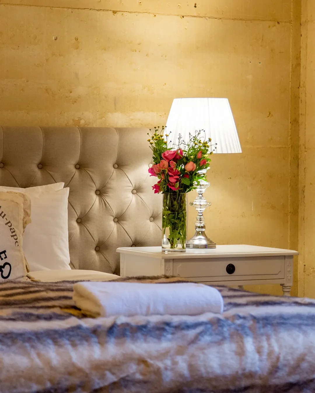 A bedroom scene with a bed having a tufted beige headboard, white pillows, and a striped blanket. Next to the bed is a white nightstand with a glass vase of pink and purple flowers and a glass table lamp with a white shade. The background features a yellow wall.