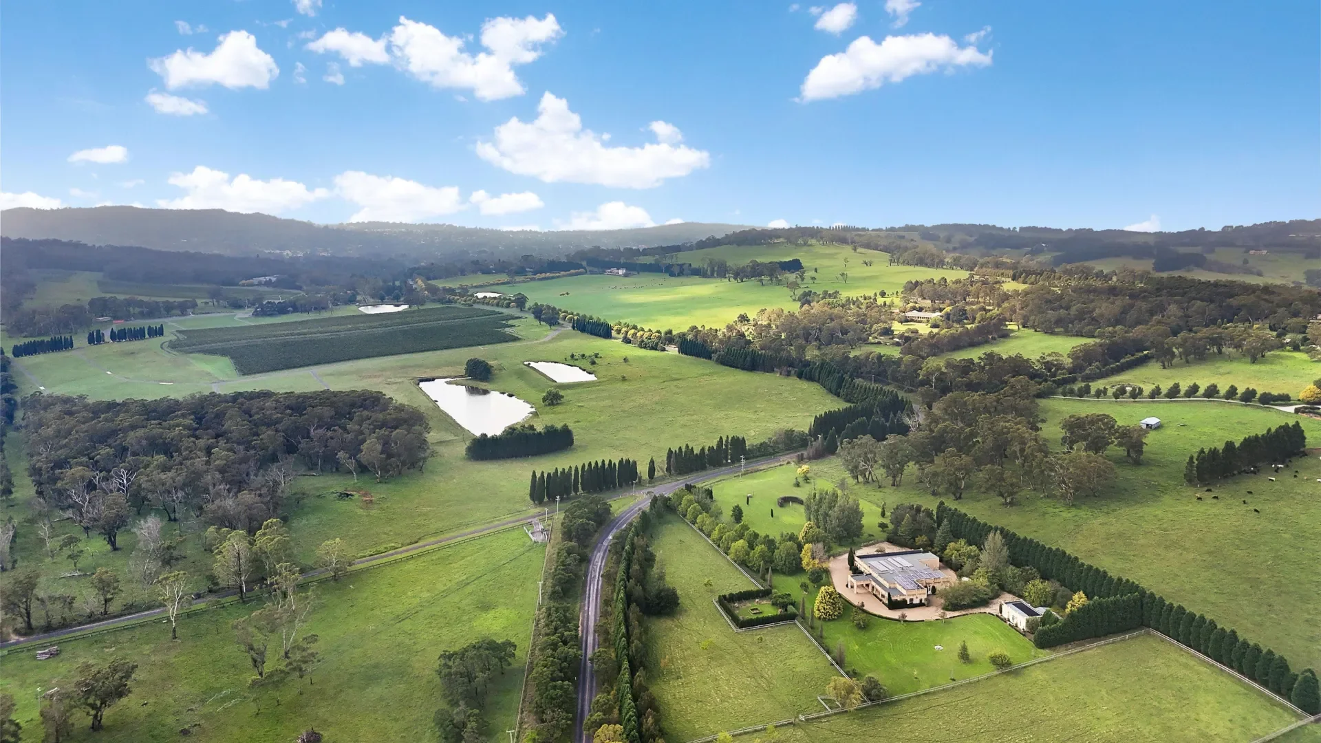 Aerial view of a pastoral landscape with green fields, trees, small ponds, and a few buildings under a partly cloudy sky.