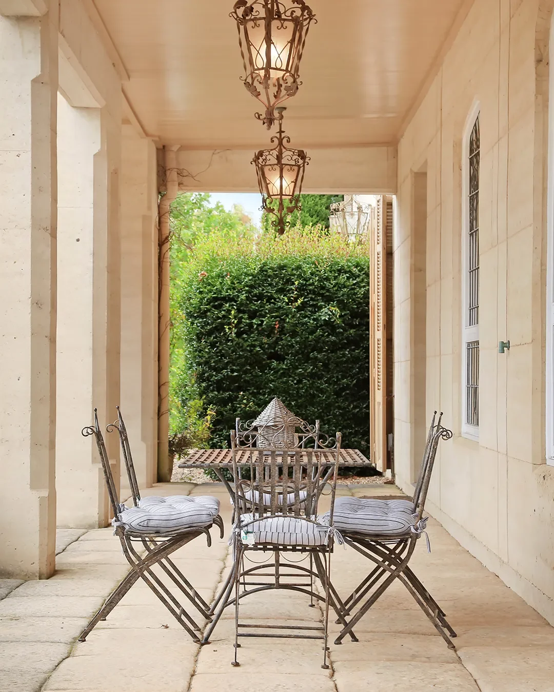 Outdoor patio with metal dining table and four chairs with striped cushions, hanging lanterns, and lush green bushes in the background.