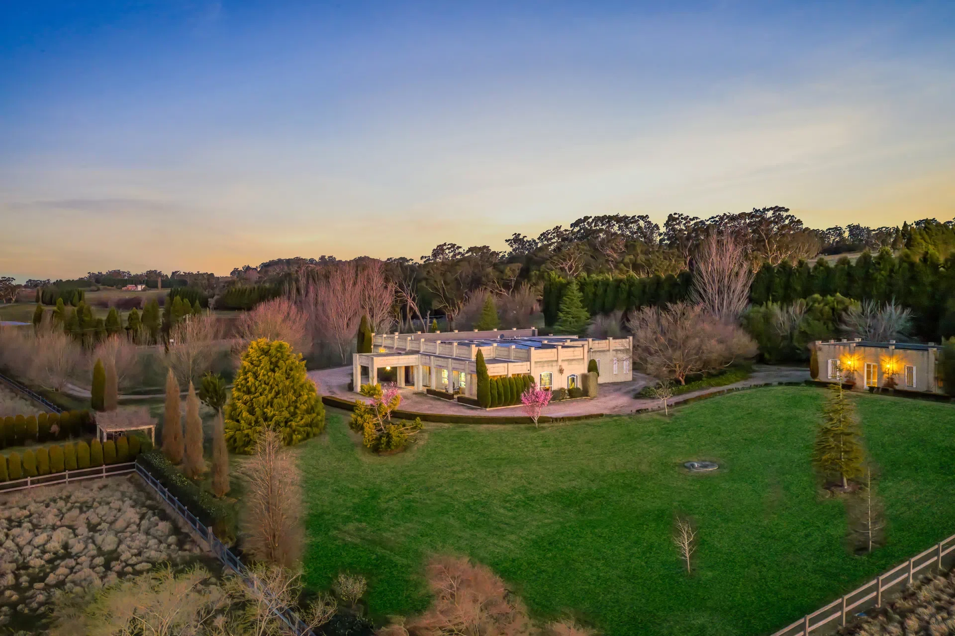 An aerial view of a large estate at sunset, featuring modern buildings, landscaped gardens, various trees, and a fenced yard, with a wooded hillside in the background.