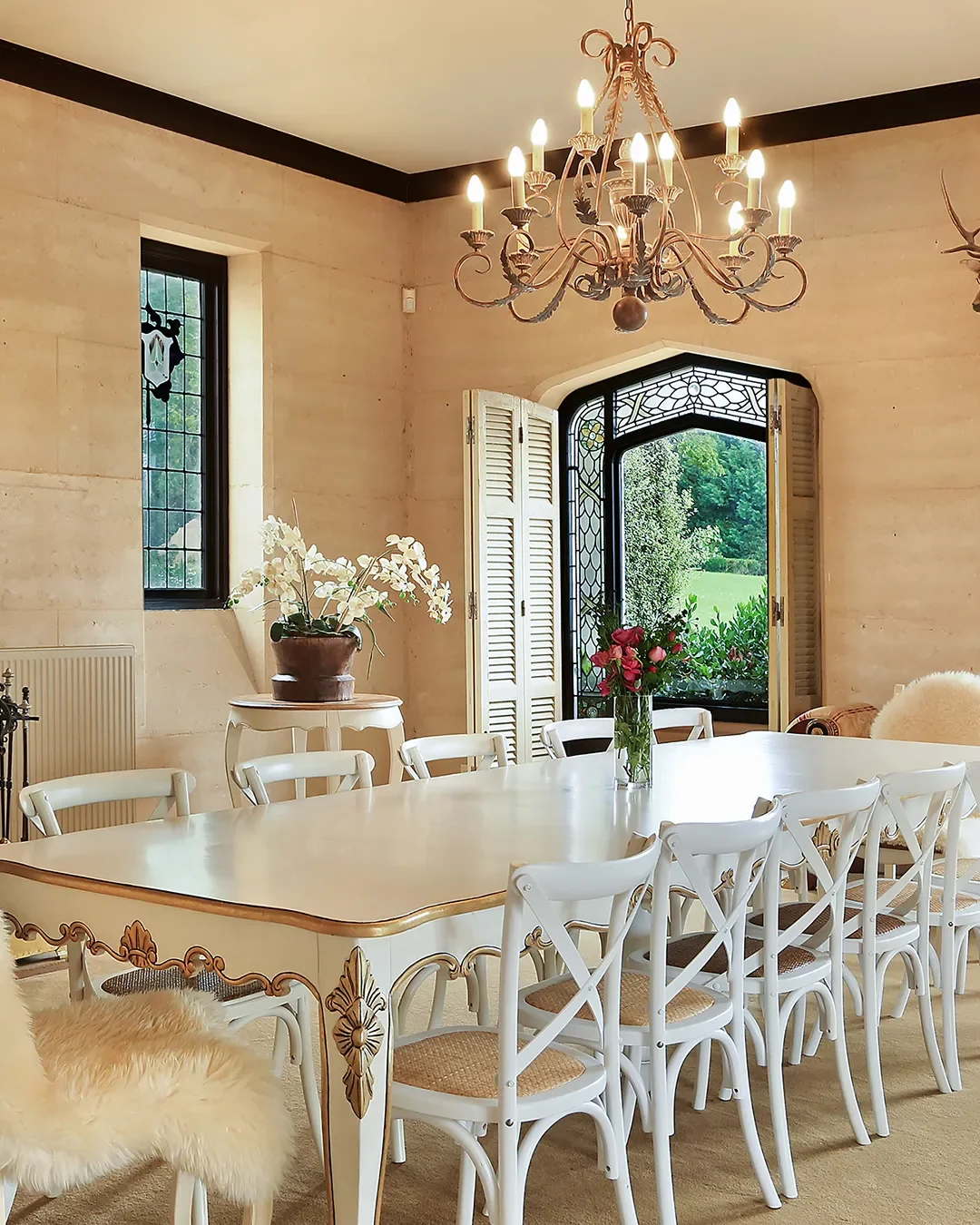 Elegant dining room with a large white table surrounded by white chairs with woven seats, a chandelier hanging from the ceiling, a window and a open door with shutters, and potted plants and flowers for decoration.