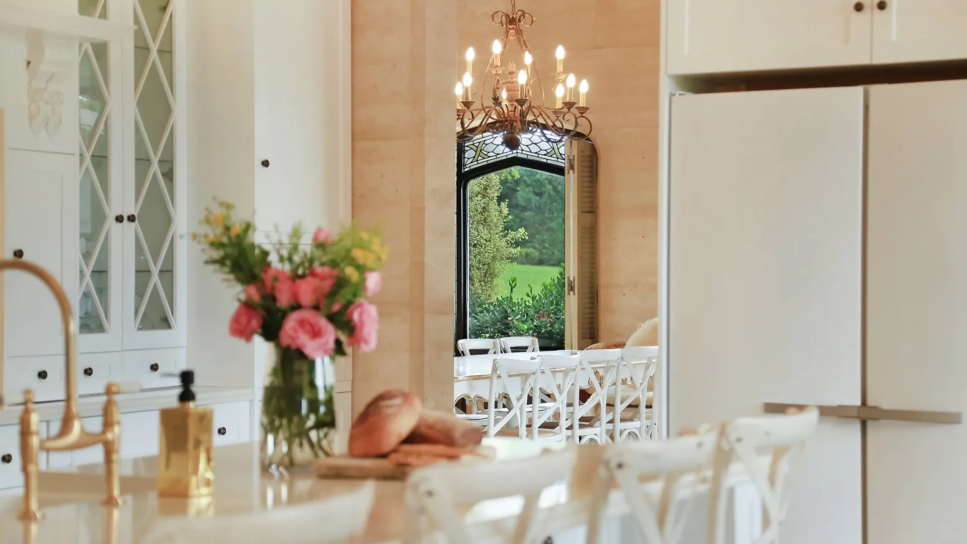 A bright dining room with white chairs around a table, a chandelier hanging from the ceiling, a window with a view of greenery outside, a bouquet of pink flowers in a vase on the table, and a white cabinet with glass doors.