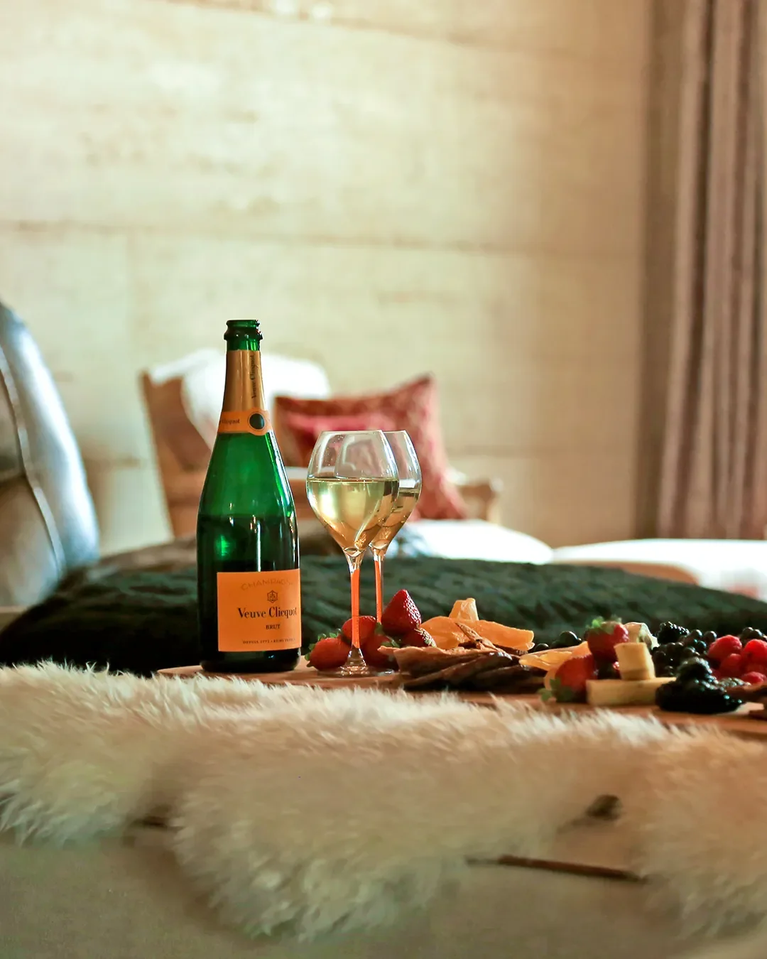 A table with a champagne bottle, two glasses of white wine, a variety of cheese, strawberries, blackberries, and other assorted berries, set in a cozy room with a bed and pillows in the background.