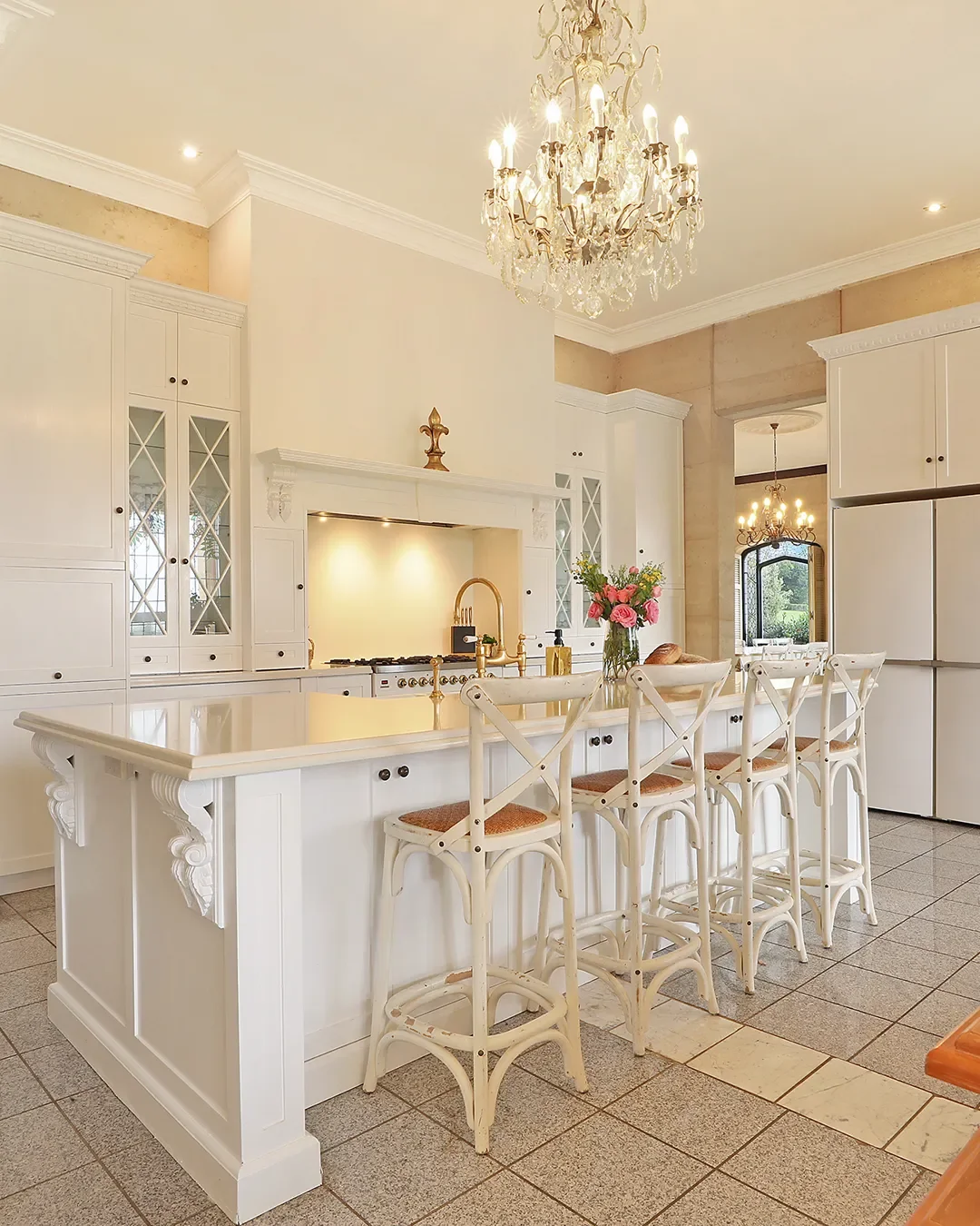 A bright, elegant kitchen with white cabinets, a large white island, and gold fixtures. A chandelier hangs from the ceiling, and four white barstools are lined up at the island. A vase of pink flowers and a bread loaf sit on the countertop.