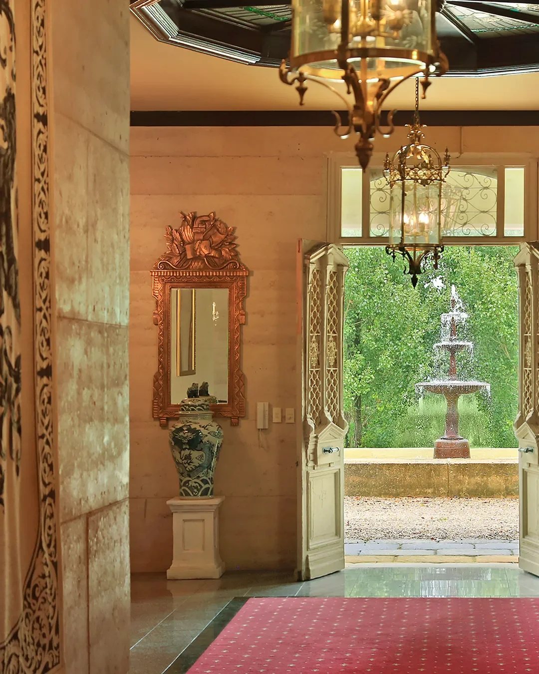 Entrance hall with open double doors leading outside to a stone courtyard and fountain, ornate chandelier, mirror with decorative frame, and a large vase on a pedestal.