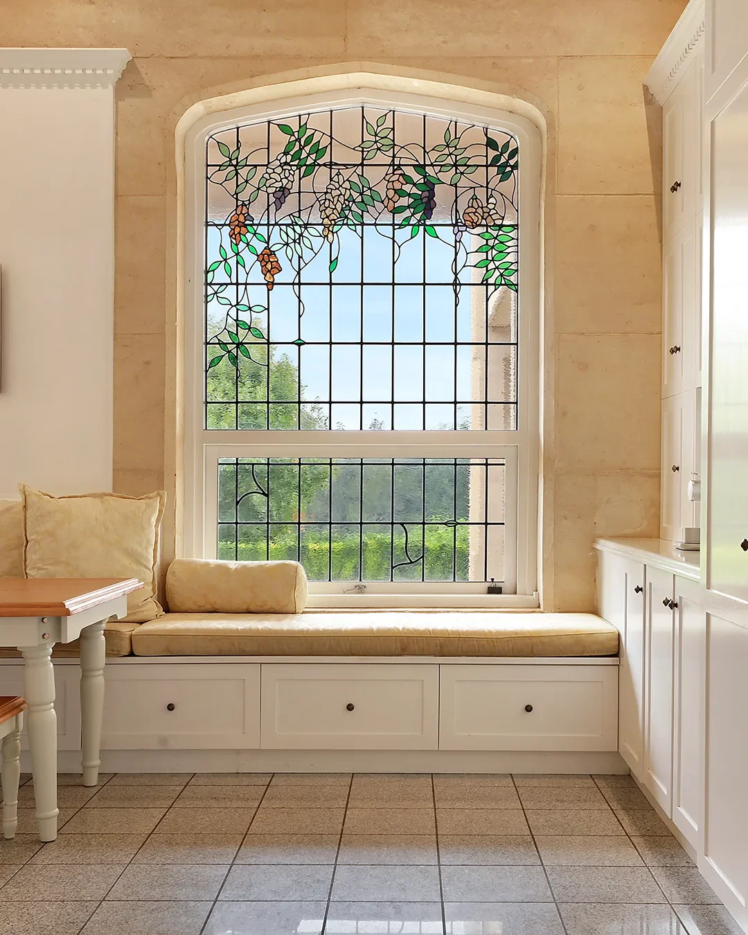 A stained glass window with a grapevine design, featuring green and purple vines, above a window seat with cream-colored cushions and a view of green trees outside.
