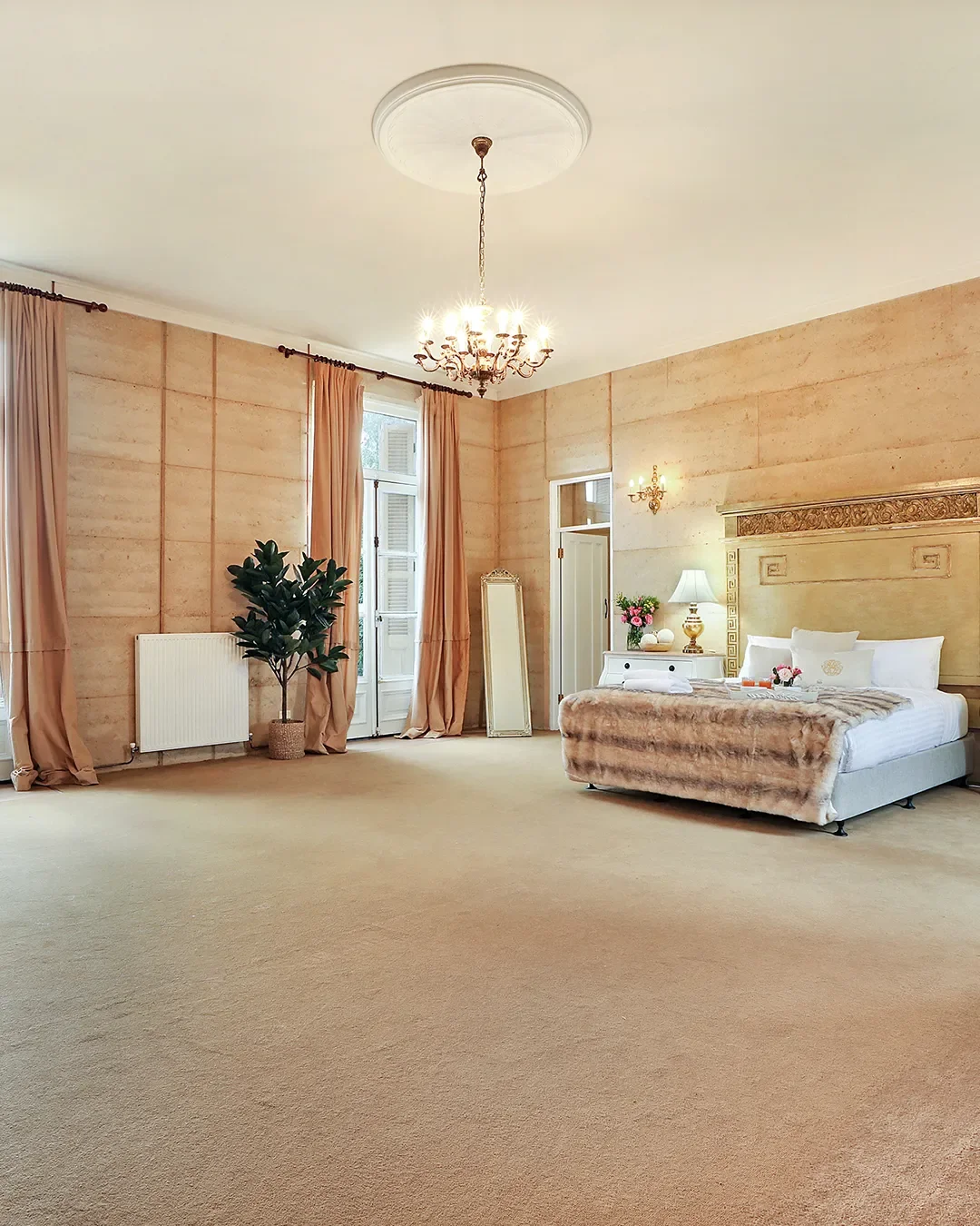 Spacious bedroom with beige walls, pink curtains, a chandelier, a large bed with white linens and a textured blanket, a white bedside table with a lamp, flowers, and decorative items, a tall mirror, a potted plant, and windows with shutters.