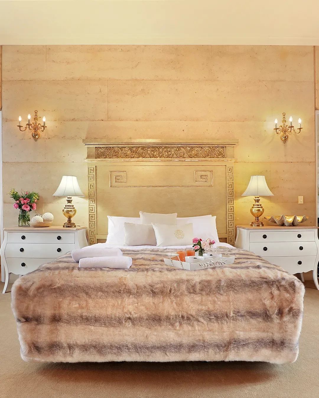 Luxurious hotel bedroom with beige walls, ornate headboard, two white nightstands with gold lamps, wall sconces, a flower arrangement, and a plush bed with white pillows and a fur blanket, featuring a breakfast tray with flowers, juice, and cups.
