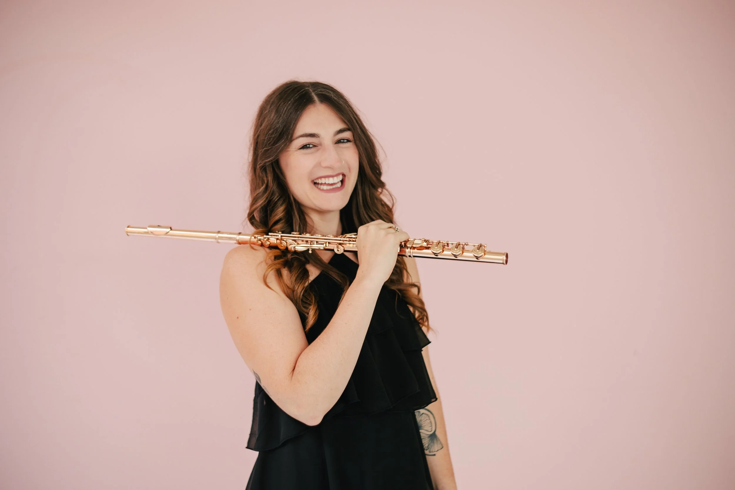 A woman, Chelsea Tanner, with long brown curly hair smiling and holding a shiny gold flute against a pink background.