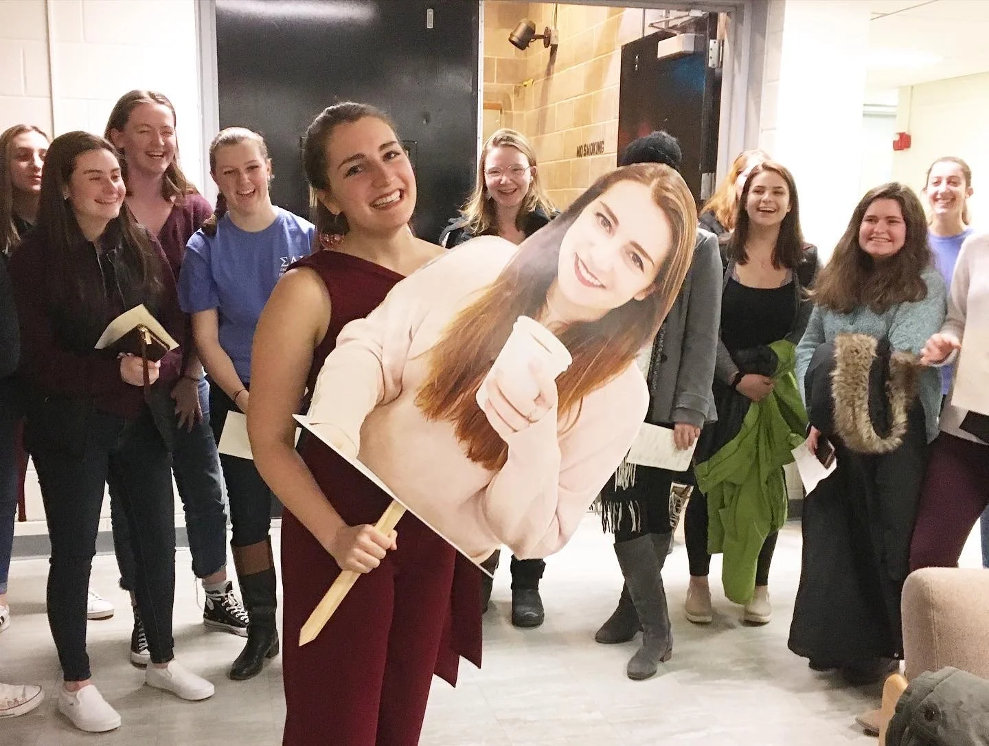 Group of young women at a social event, with one holding a large cardboard cutout of a smiling woman holding a coffee cup.