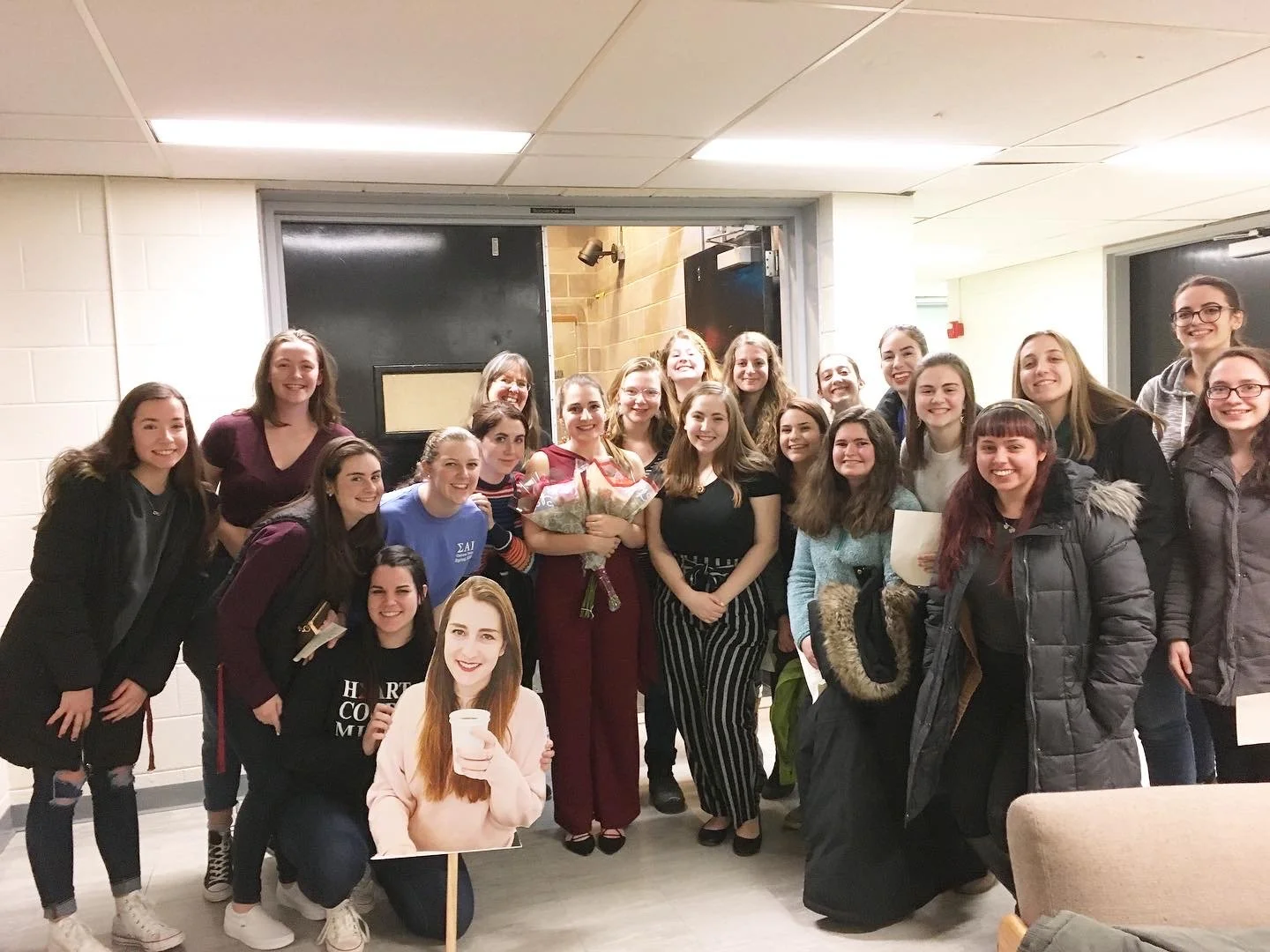 Group of women standing in a hallway, some holding flowers and a large cutout of a woman's face, celebrating together.