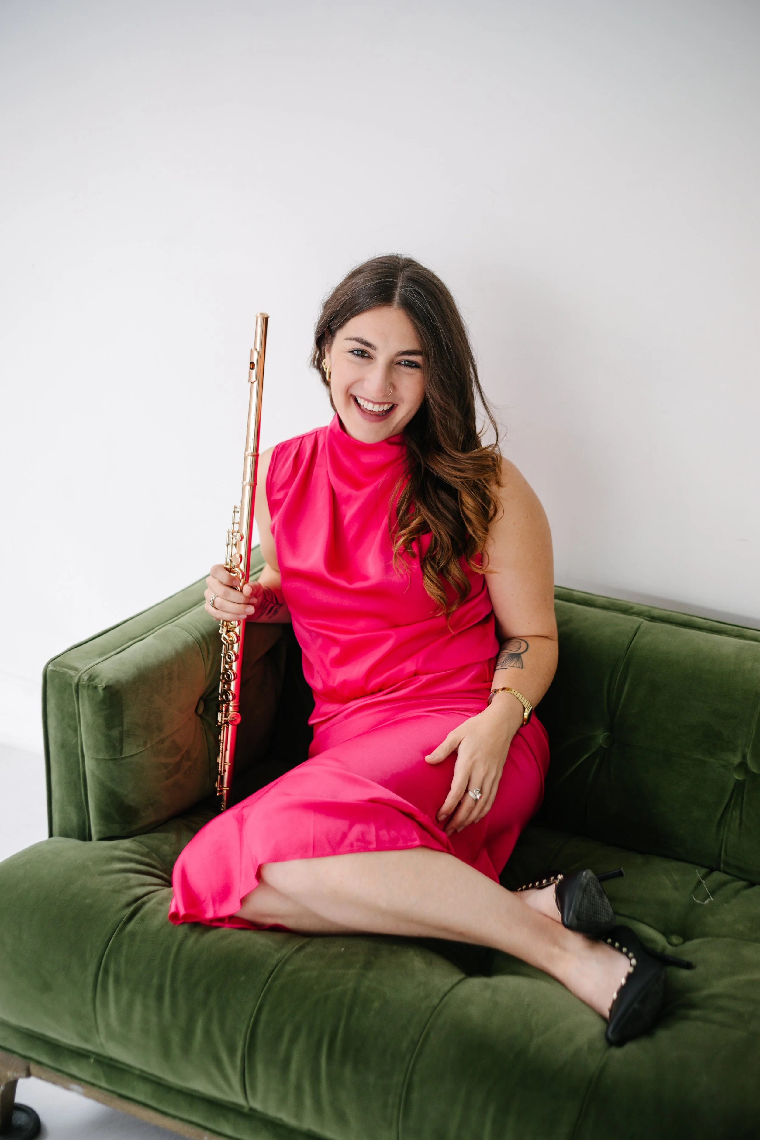 A young woman, Chelsea Tanner, with long brown hair, wearing a bright pink dress, sitting on a green velvet couch, holding a flute, laughing, and looking at the camera.