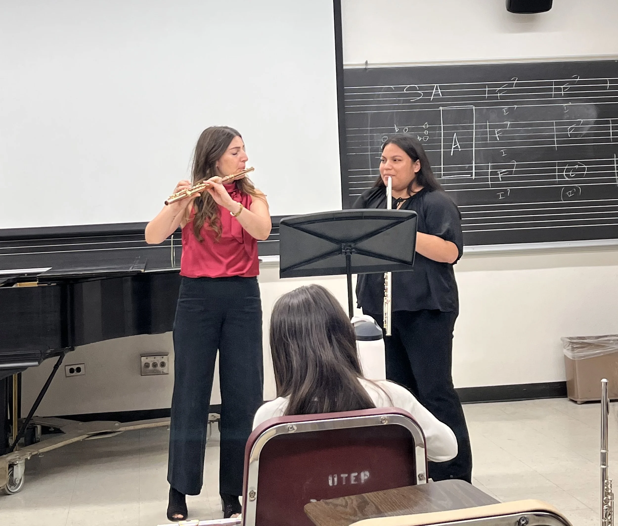 Two women in a classroom, one playing a flute and the other holding a flute near her mouth, with musical notation on the blackboard behind them. Several classroom chairs and a music stand are visible.