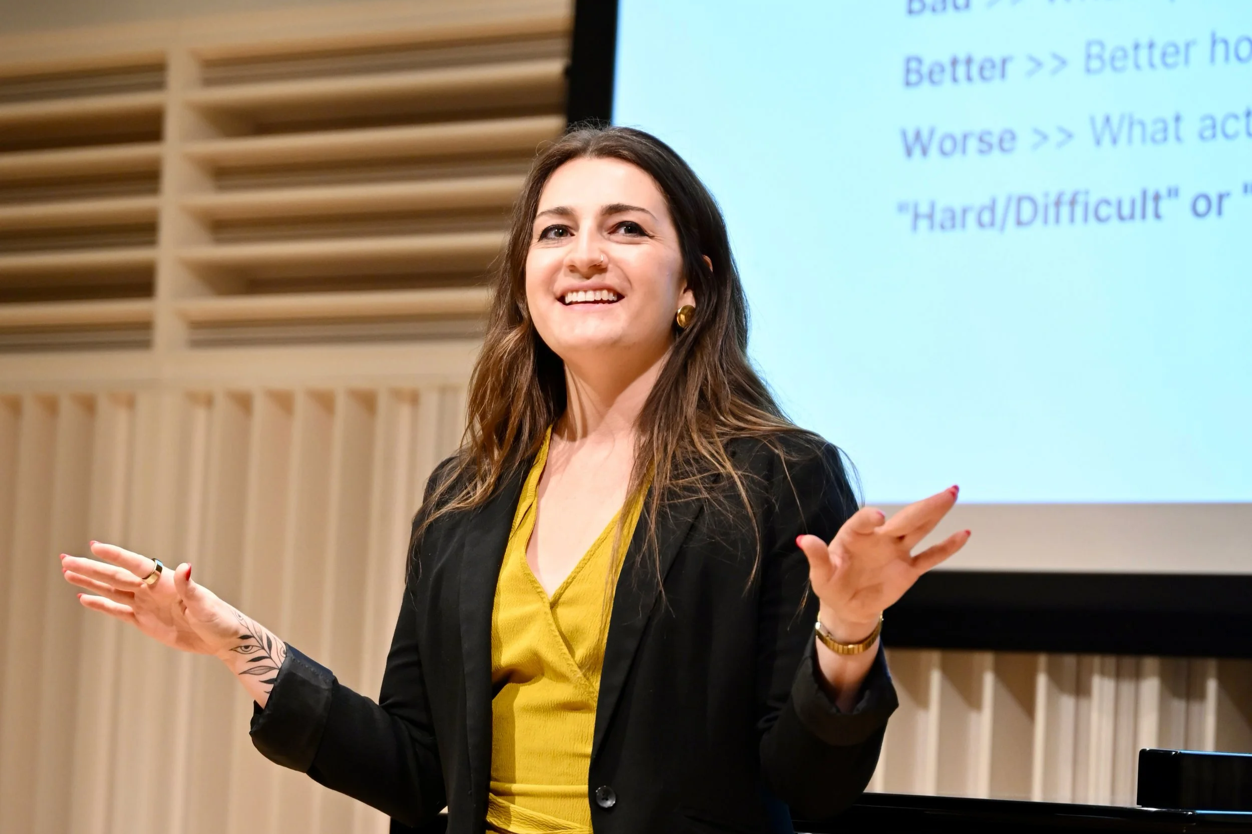 A woman with dark brown hair, wearing a black blazer and yellow top, stands in front of a presentation screen, smiling and gesturing with her hands while giving a presentation.