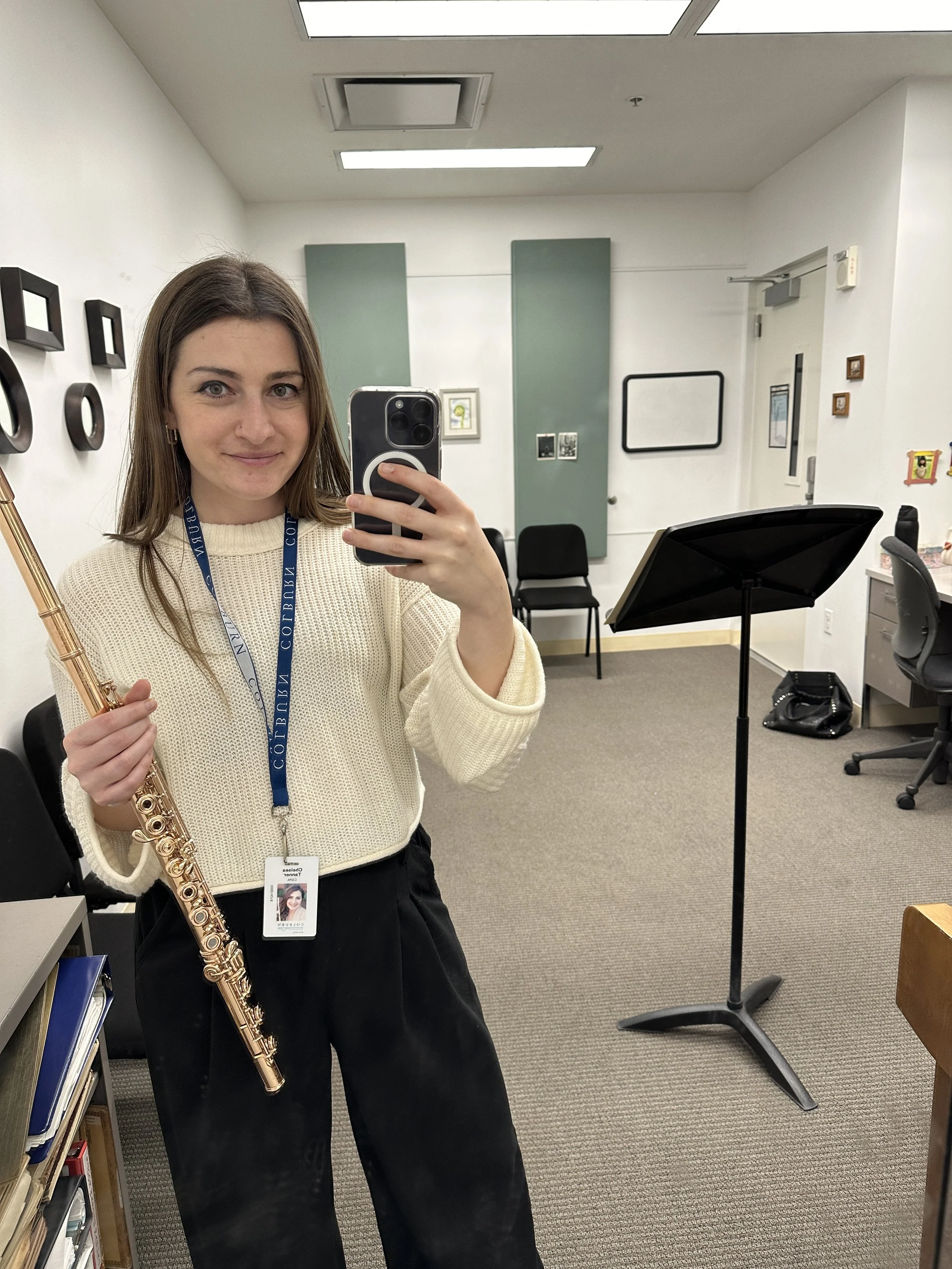 A woman is taking a selfie in an office or classroom while holding a gold flute. She wears a beige sweater, black pants, and has a college ID badge around her neck. The room has chairs, framed pictures, and a music stand.