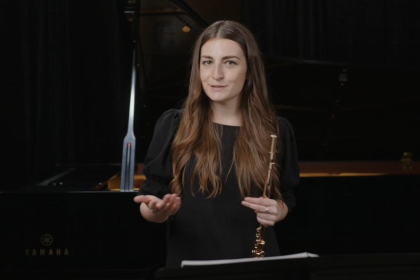 A young woman holding a musical instrument, standing in front of a grand piano indoors.