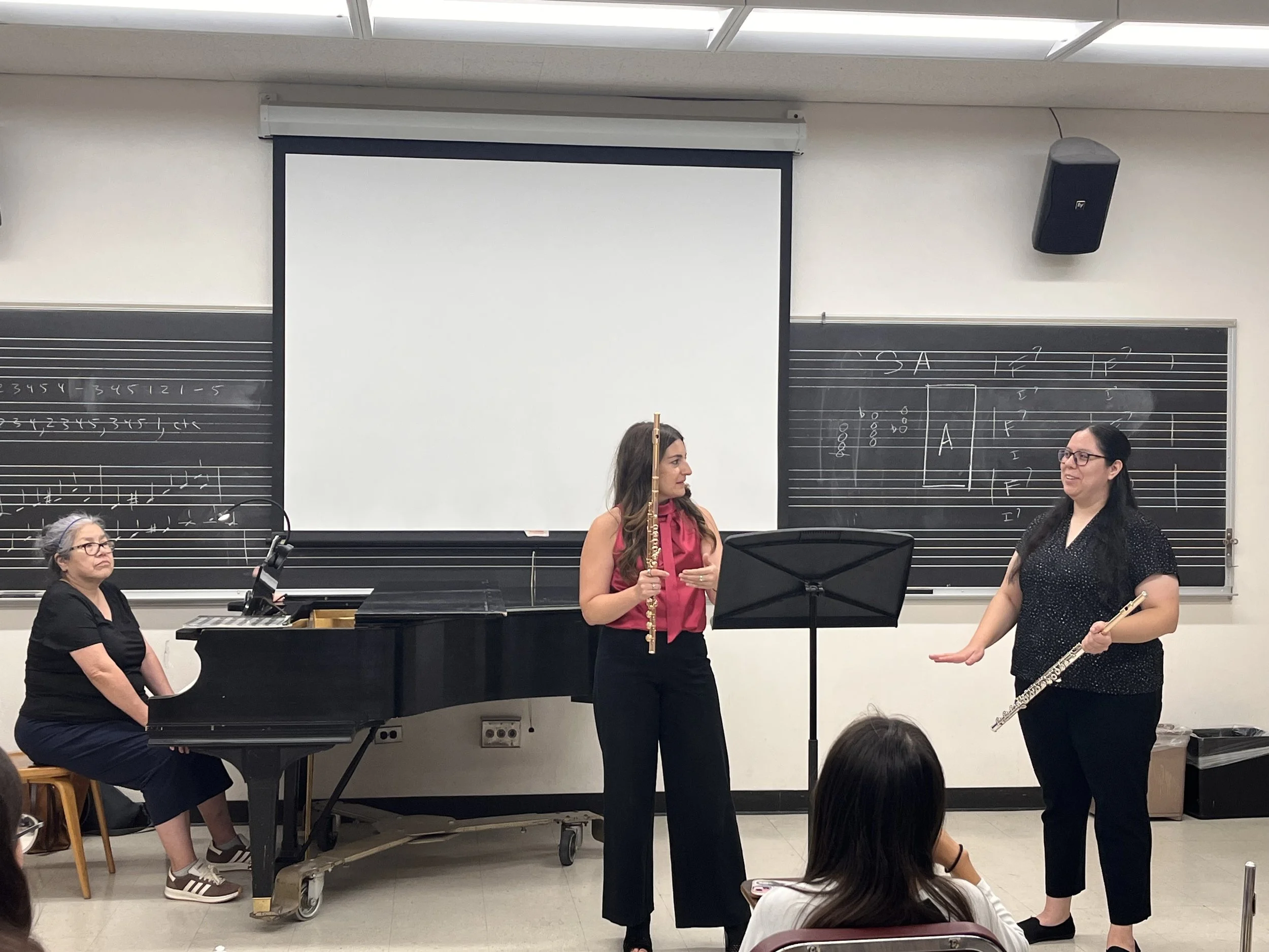 A classroom setting with women musicians performing with a piano, flute, and other instruments in front of a blackboard and projection screen.