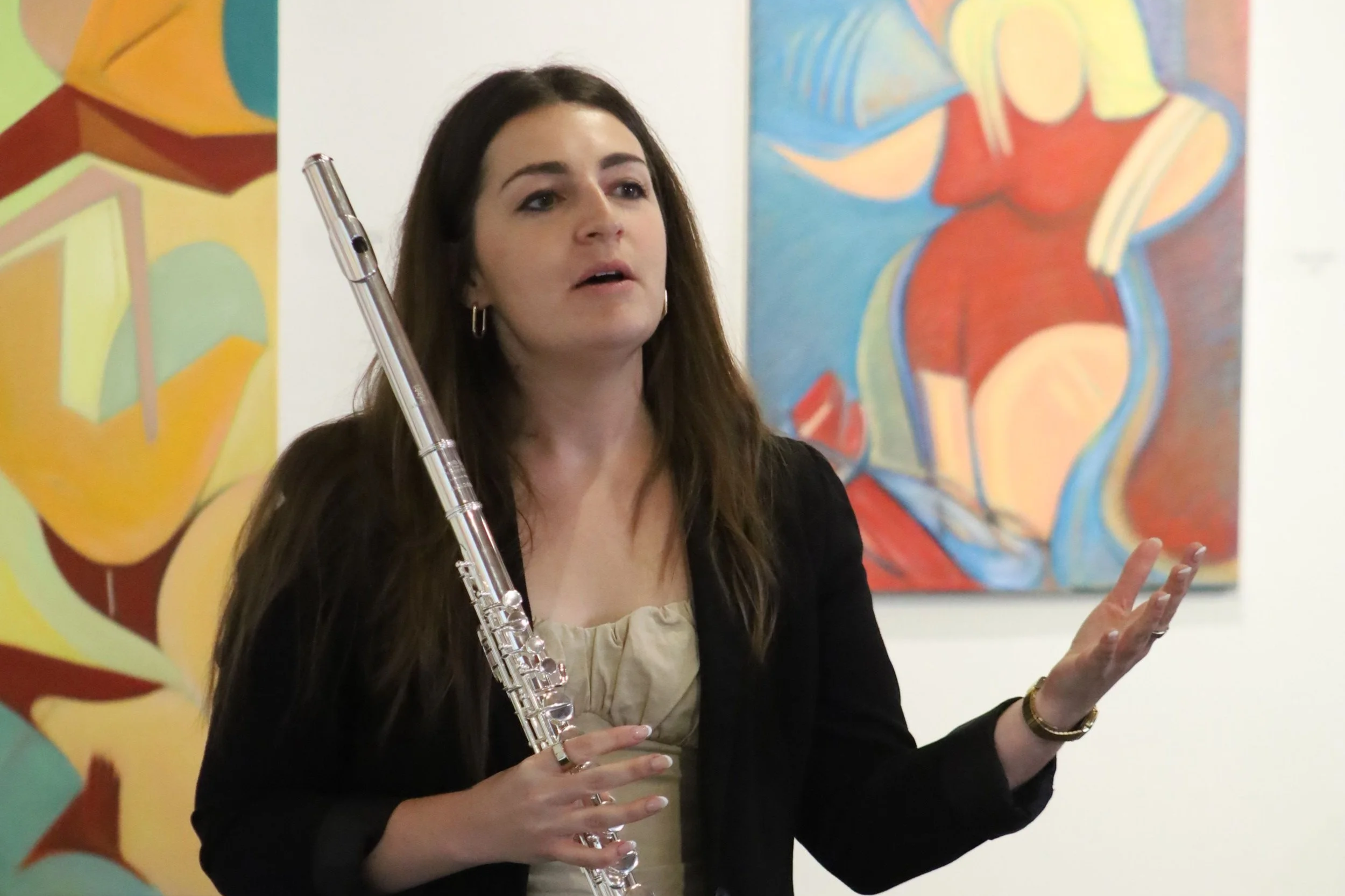 A woman playing a silver flute at an art gallery, with colorful abstract paintings in the background.
