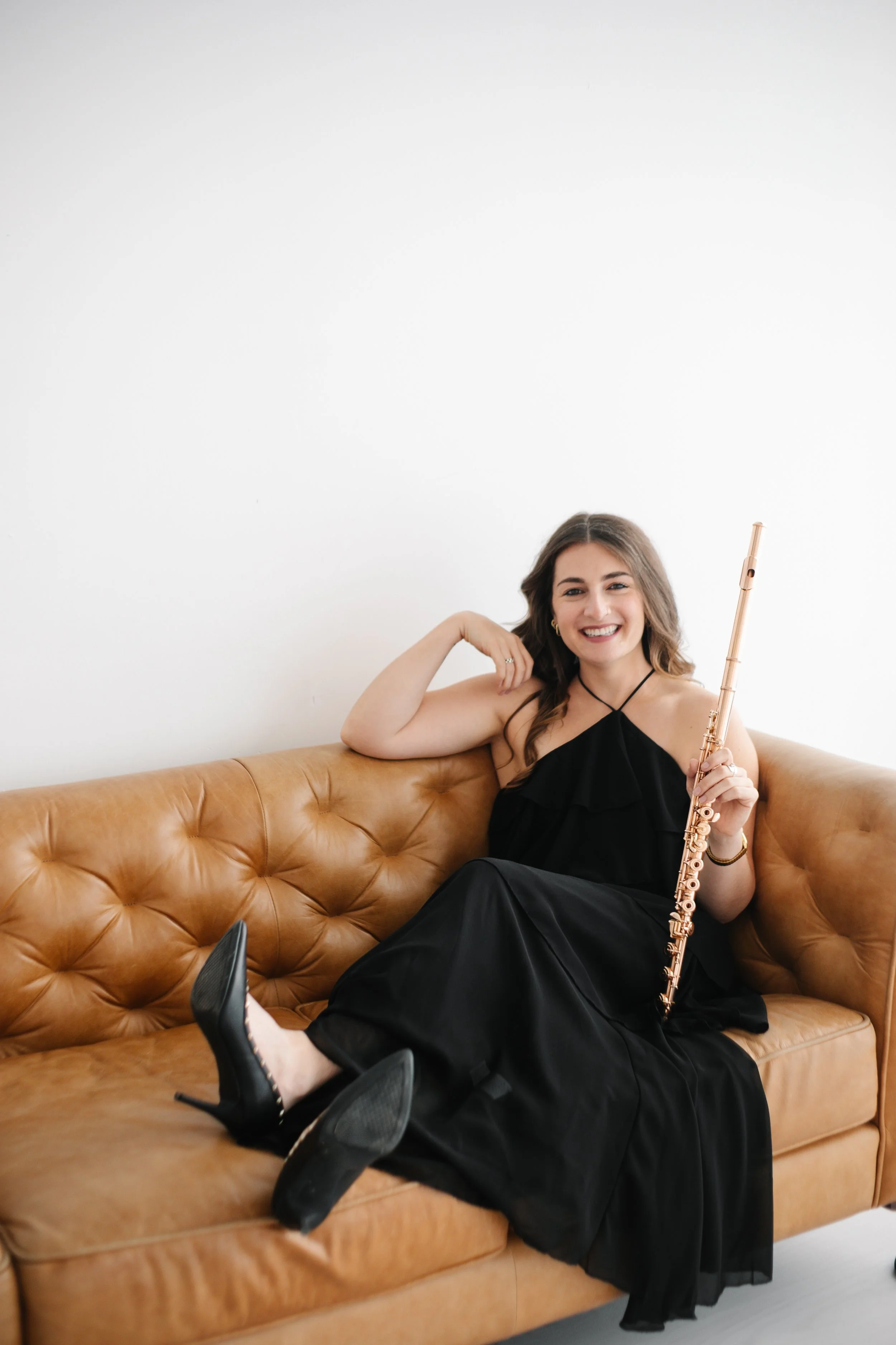 A young woman, Chelsea Tanner, with long brown hair, wearing a black dress, black high heels, and jewelry, sitting on a tan leather couch, holding a flute, and smiling at the camera.