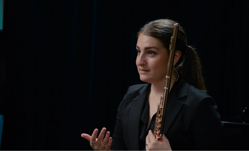 A woman in a black blazer holding a flute, sitting in a dark room with a black background.