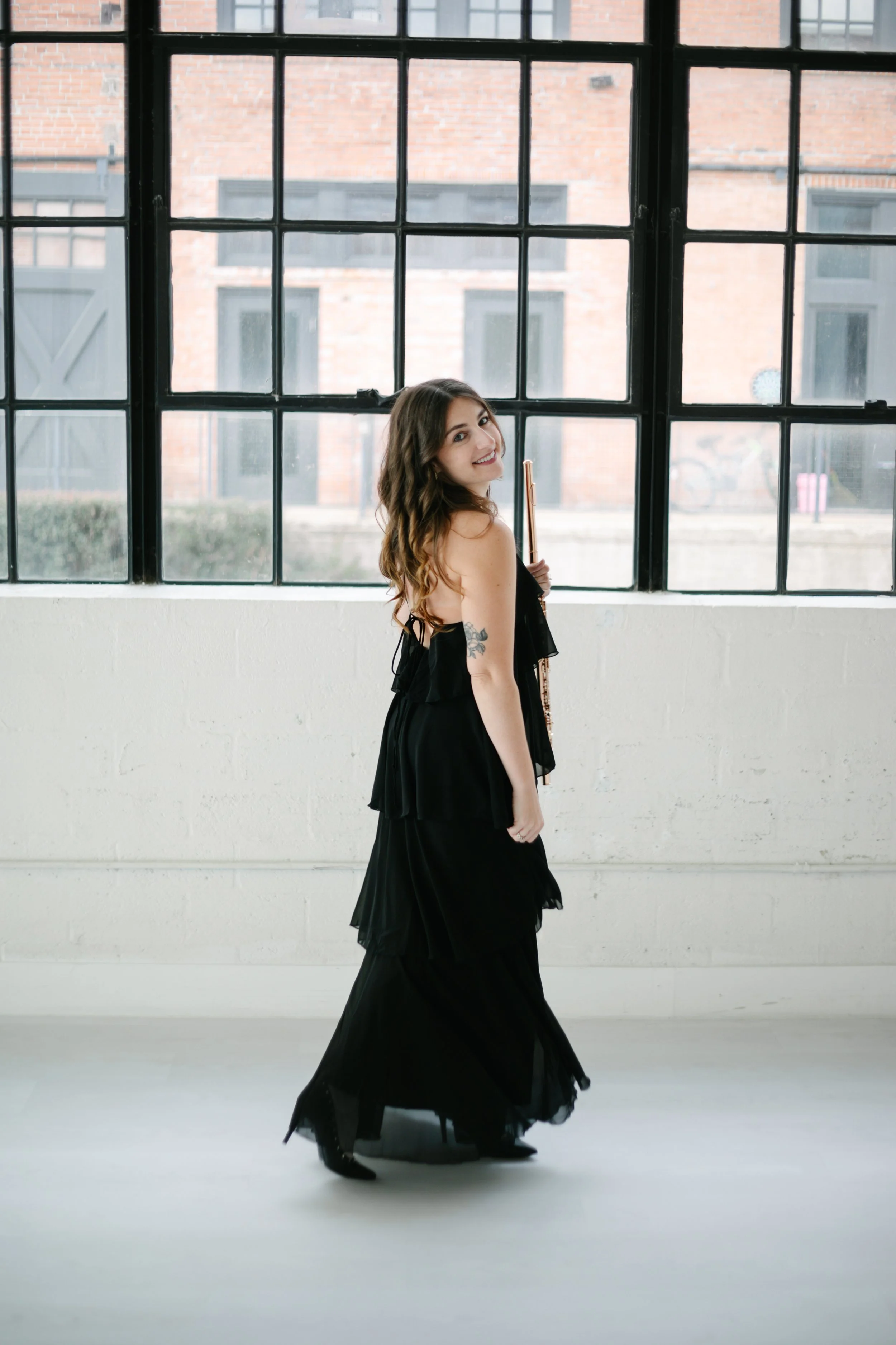 A woman , Chelsea Tanner, with long, wavy brown hair wearing a black dress and black high heels standing indoors near large window with a grid pattern, holding a long recurve bow, with exposed brick buildings visible outside.