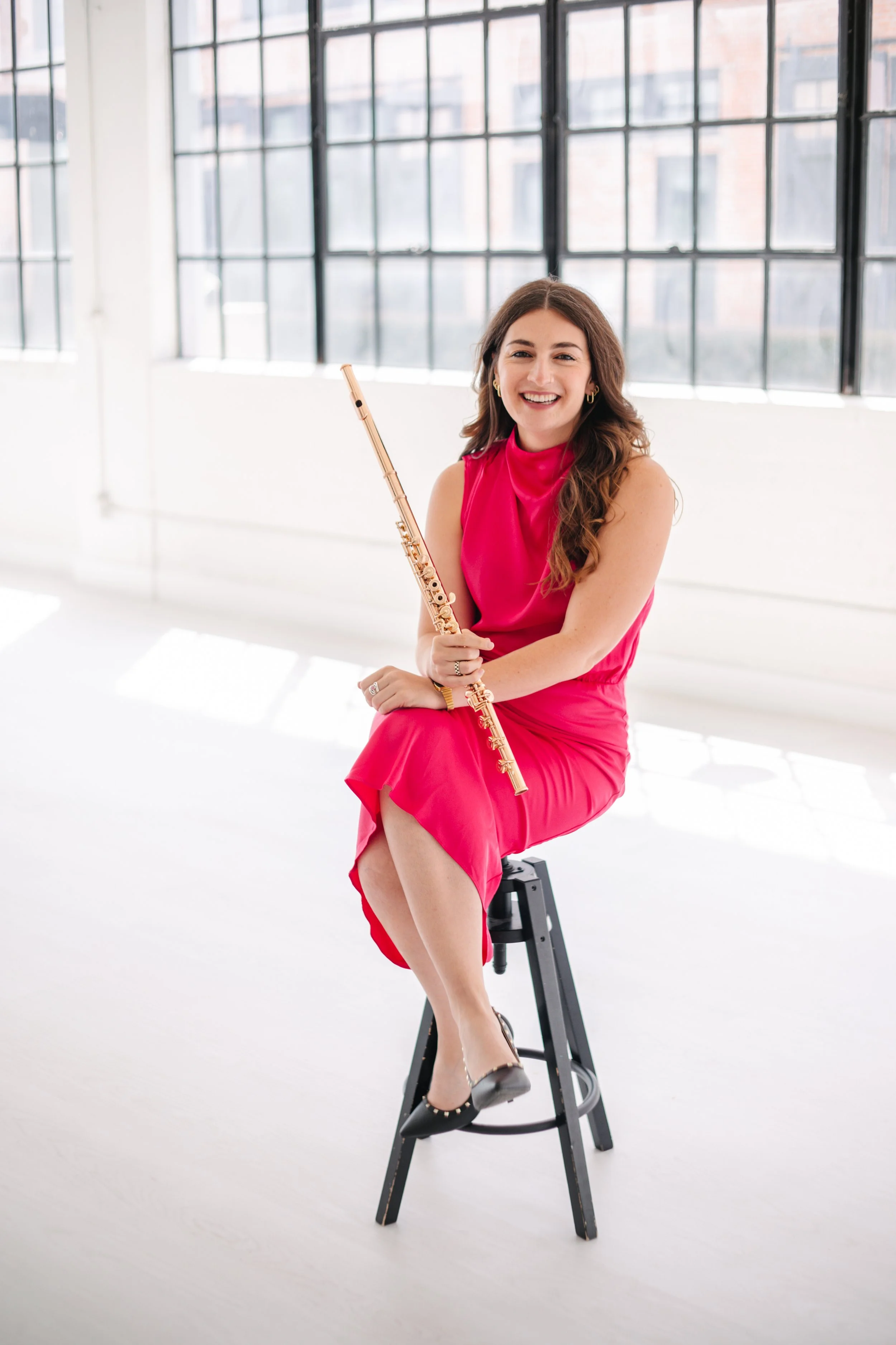 A woman, Chelsea Tanner, in a pink dress sitting on a black stool in a bright room holding a flute, with large windows in the background.