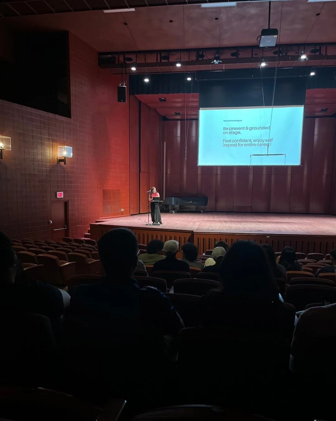A woman standing at a podium on a stage delivering a presentation to an audience in a large auditorium with wooden walls. A large screen displays presentation slides.