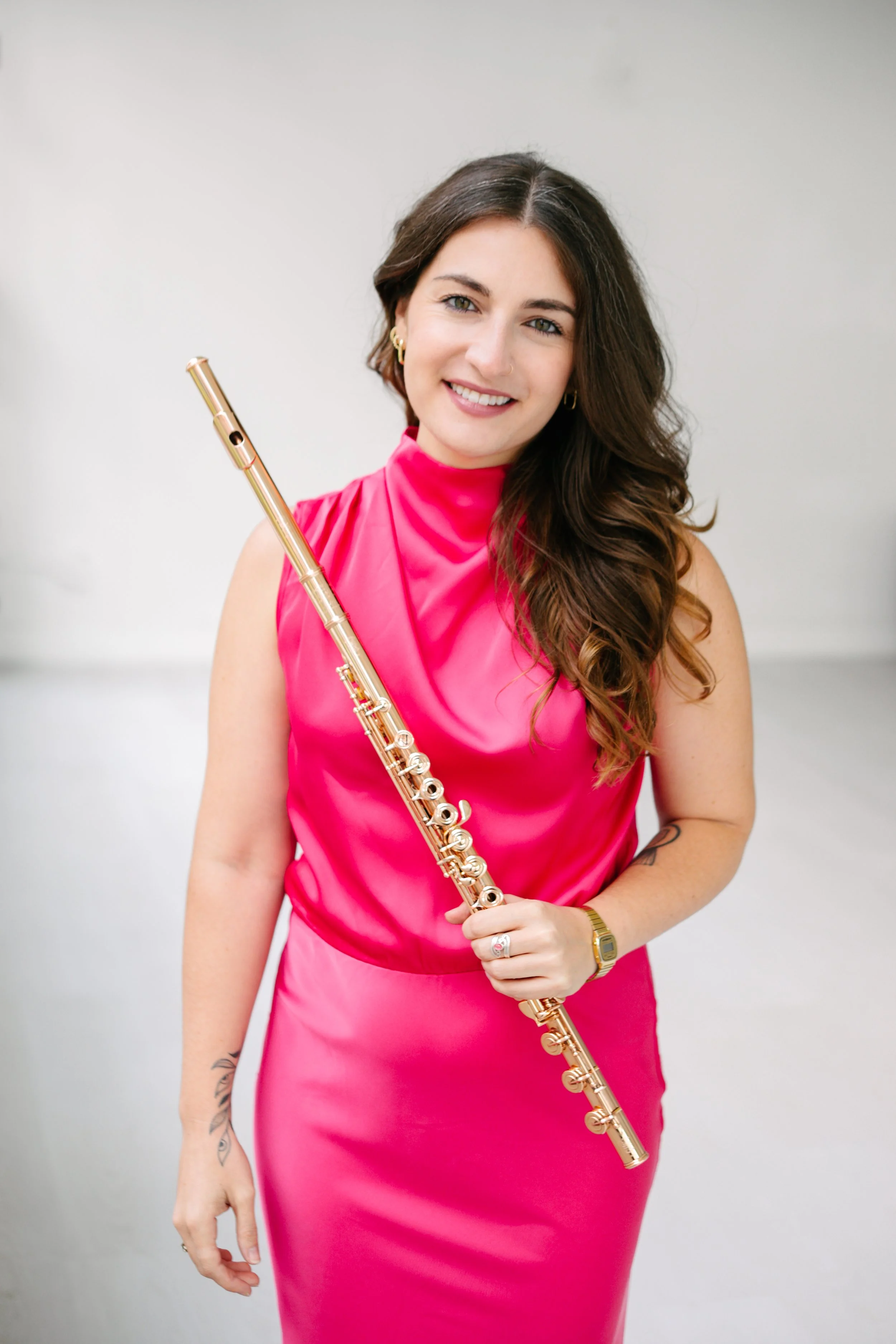 A young woman, Chelsea Tanner, with long, wavy brown hair and green eyes, dressed in a bright pink sleeveless top, holding a gold flute, standing against a white background, smiling at the camera.