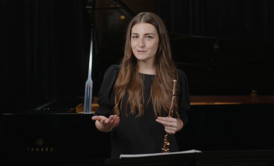 A woman holding a flute standing in front of a grand piano in a music studio or performance space.