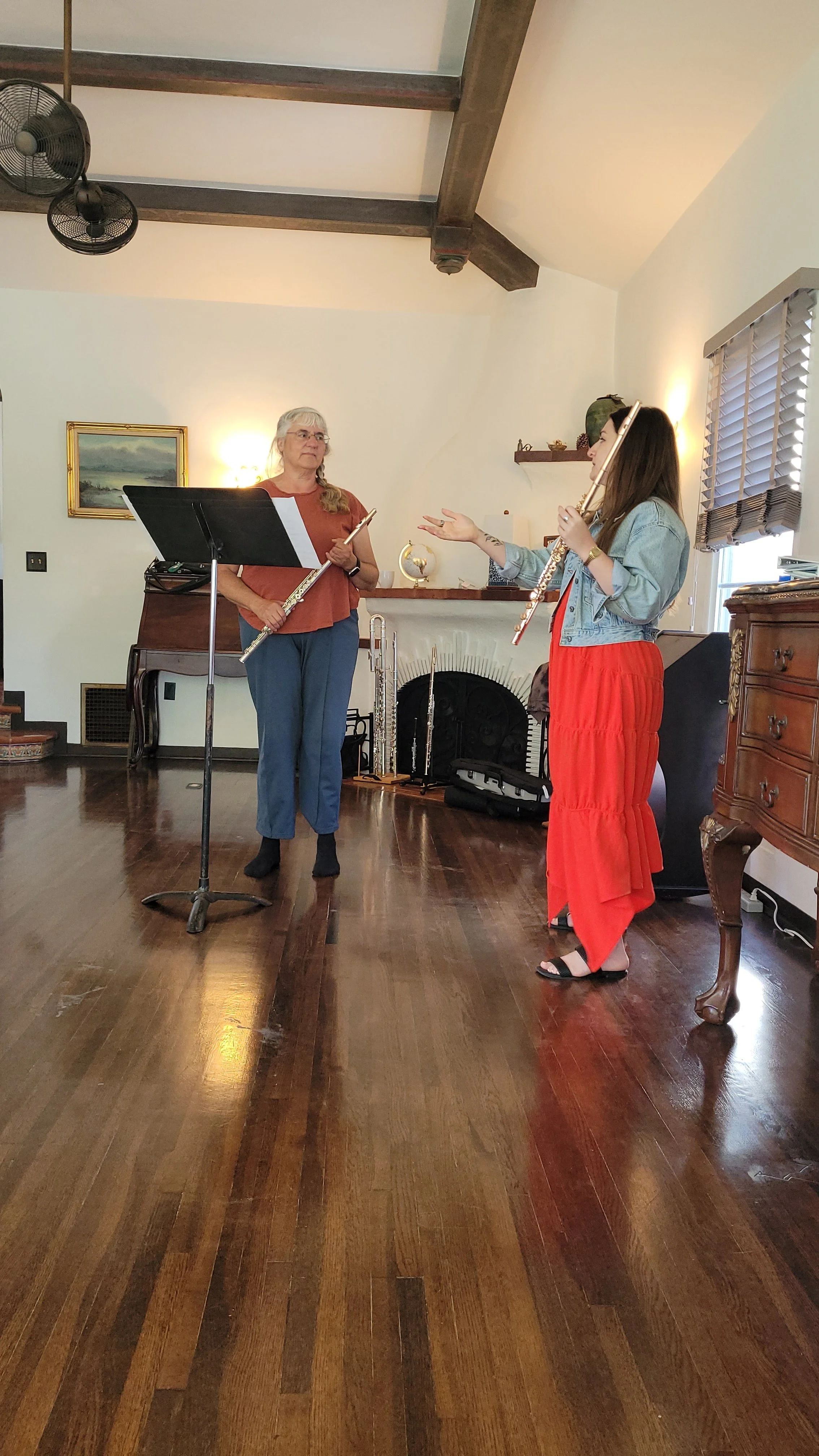 Two women indoors, one holding a music stand with sheet music and a flute, the other gesturing while holding a flute. The room has hardwood floors, a fireplace, a painting, and a window with blinds.
