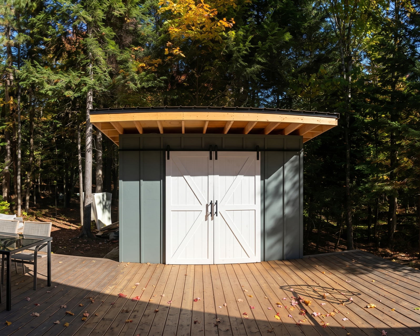 A shed with white barn-style doors and black hardware, set on a wooden deck in a wooded area with tall trees and autumn leaves.