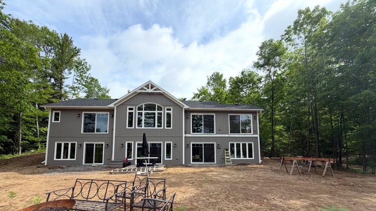 A large gray, two-story house under construction, with multiple windows, surrounded by trees, and an unfinished yard with construction materials and outdoor furniture.