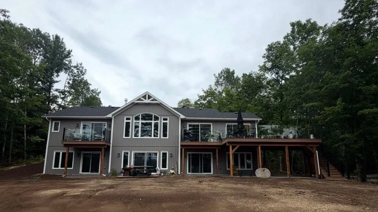 A large modern gray two-story house with multiple windows and a deck, surrounded by trees and a dirt yard in the foreground.