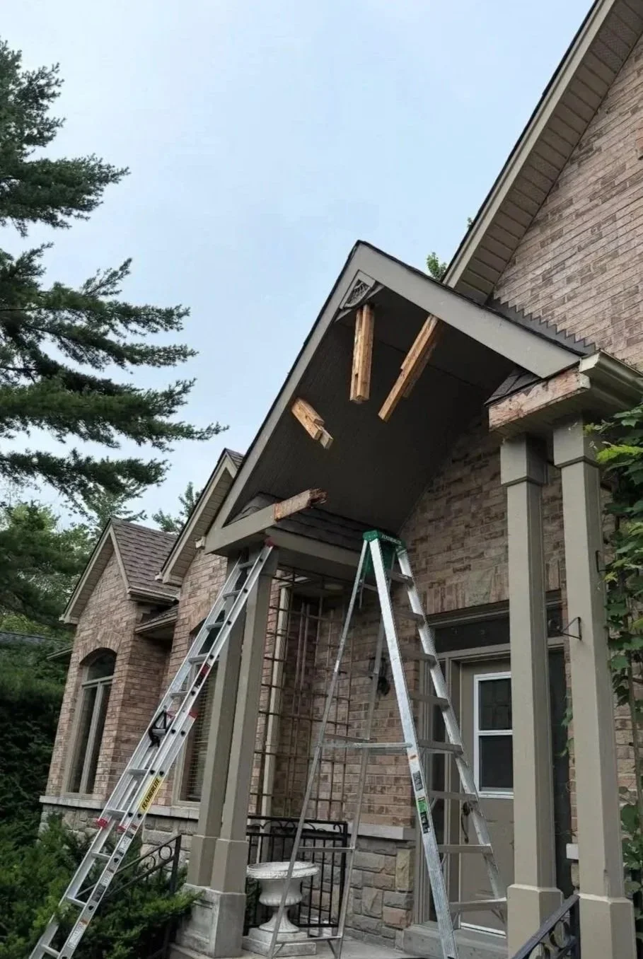 Ladders and a partially disassembled porch roof on a brick house, with trees in the background.