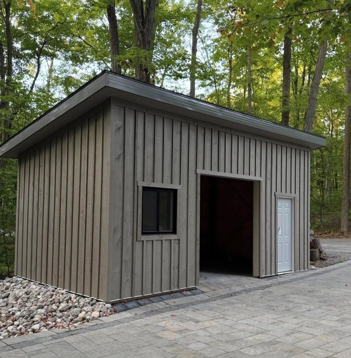 Small wooden building with a sloped roof, a window, a door, and a paved area in front, surrounded by trees.