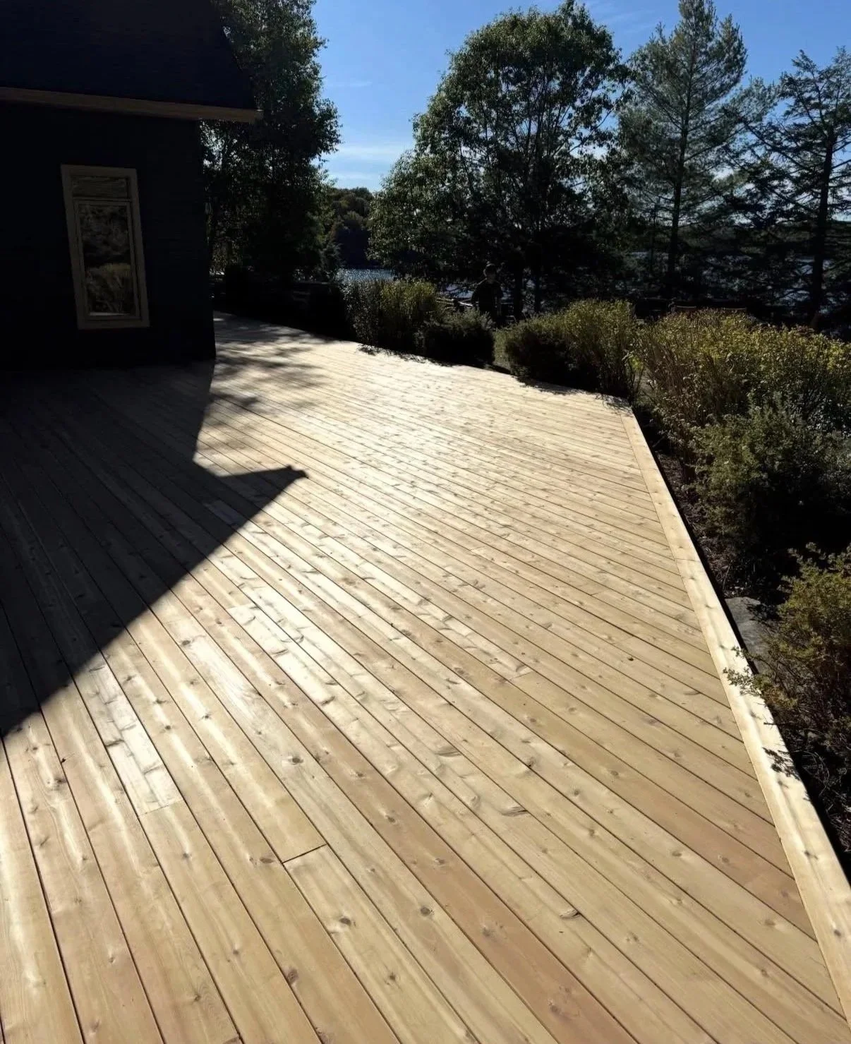 Wooden deck overlooking trees and water, with a house visible on the left side and a person in the distance.