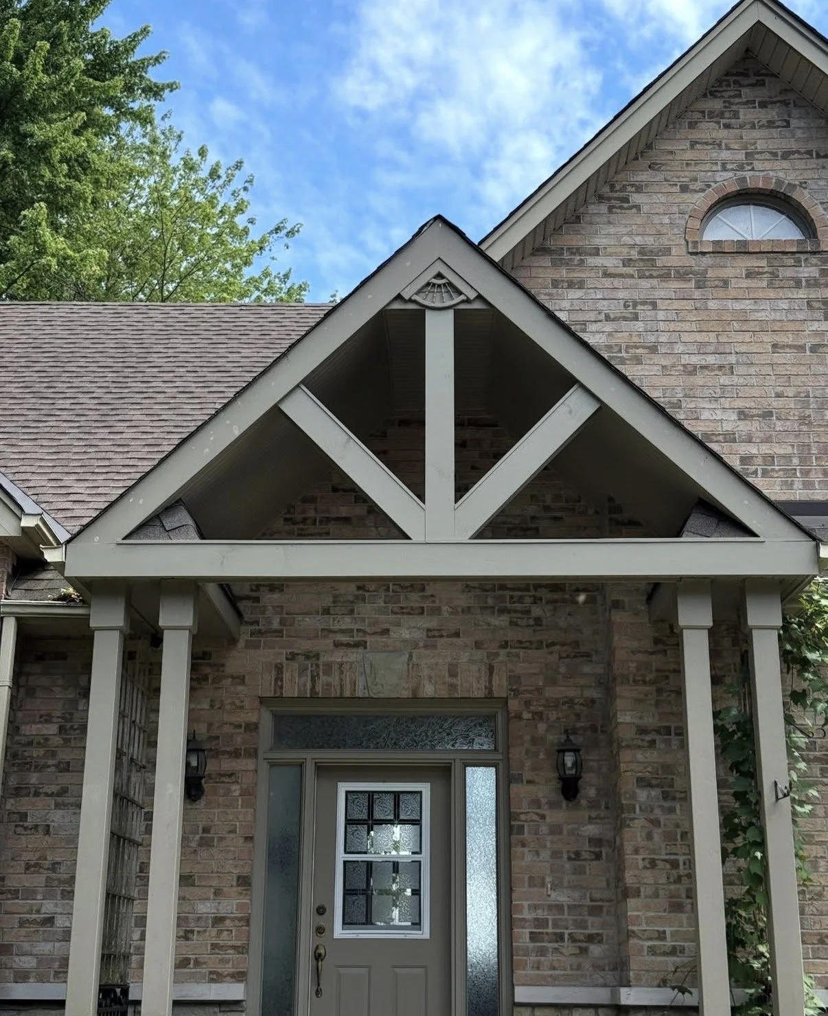 A house with a brick exterior, front door with glass window, and a gable roof with a decorative wood truss and trim, surrounded by trees and a blue sky.
