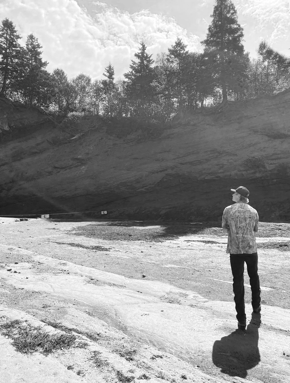 A person stands on a dirt path near a water body, looking at a cliff with trees on top. The photo is in black and white.