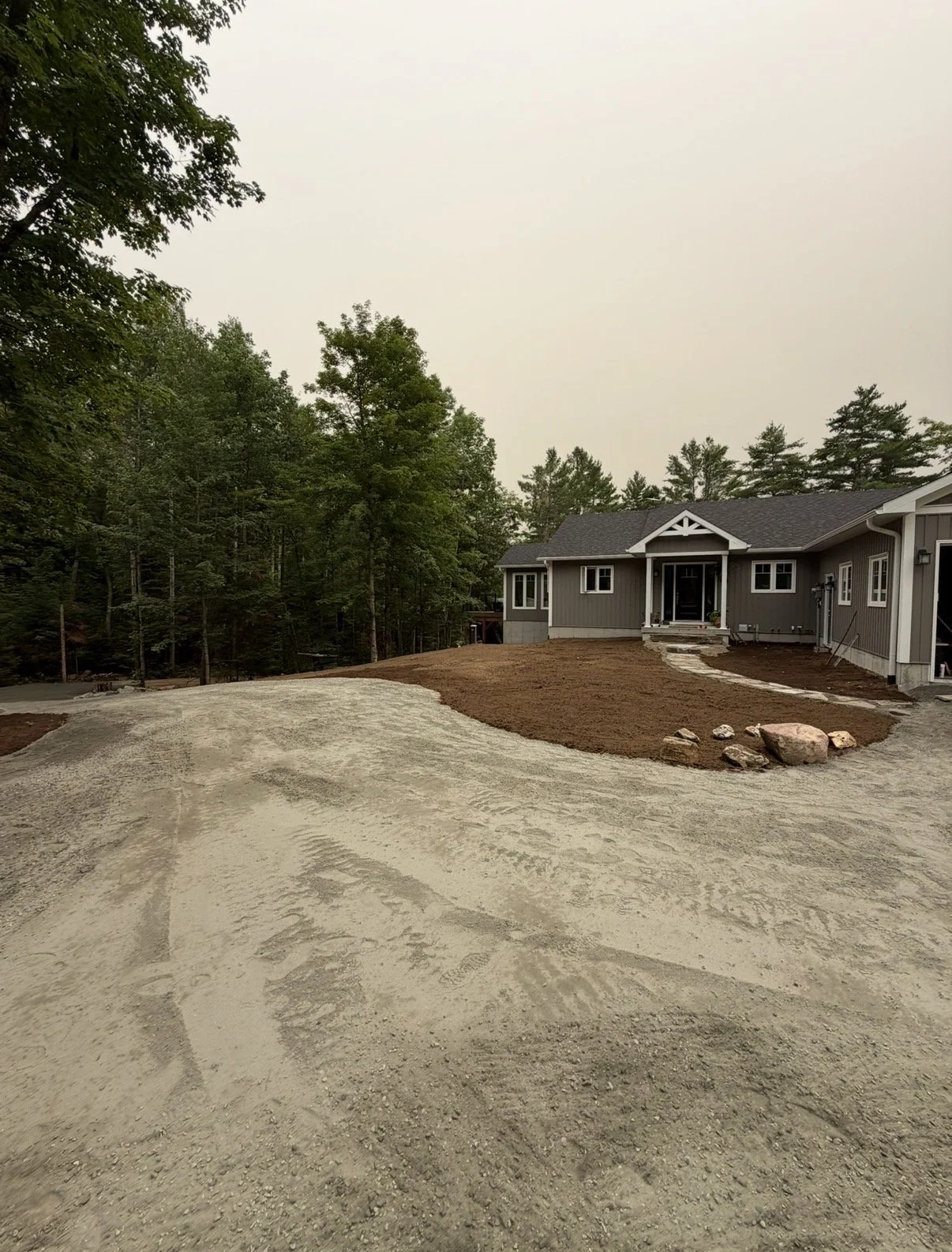 Newly constructed house with a dirt yard and unpaved driveway, surrounded by trees.