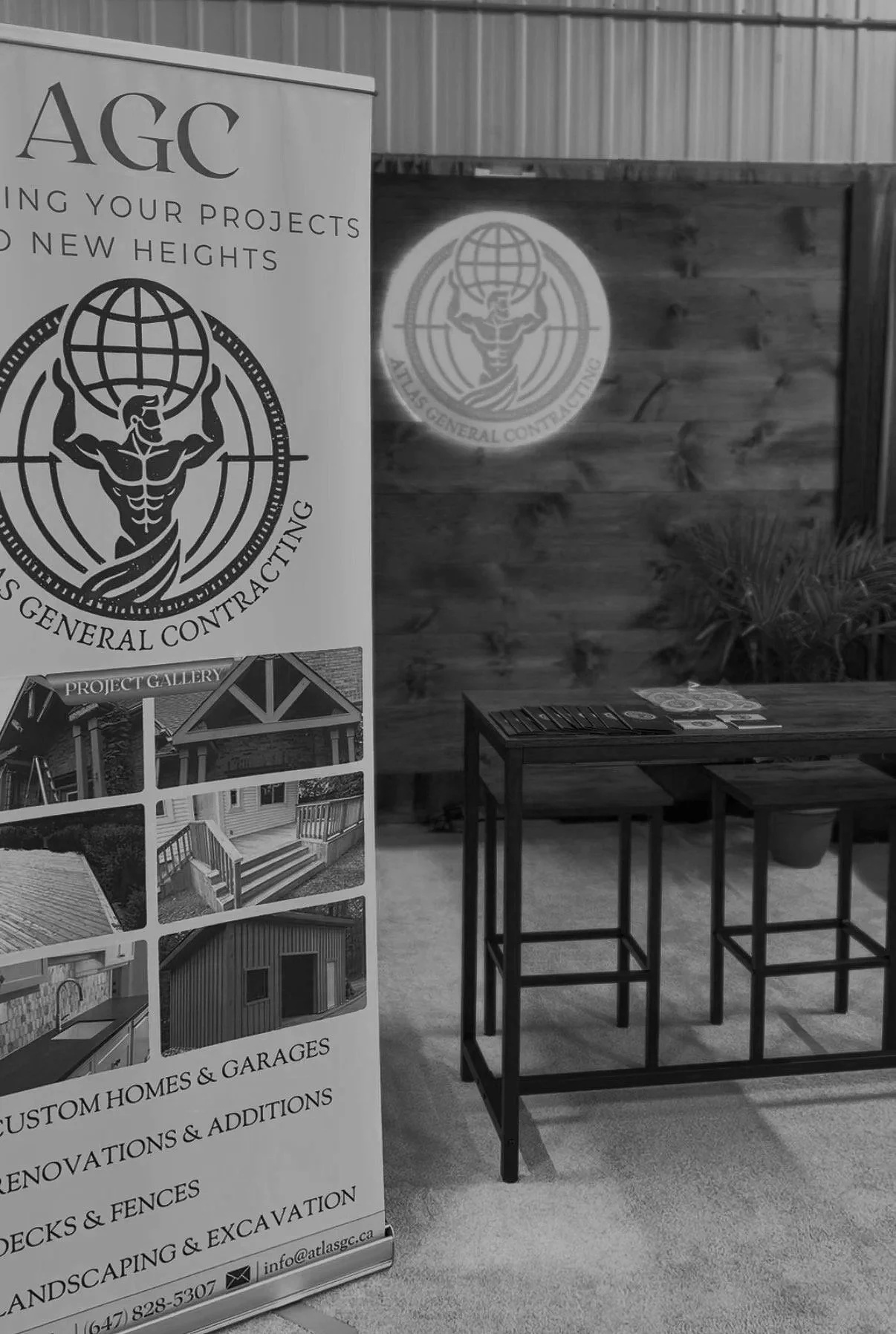 A booth with a banner displaying a logo of Atlas General Contracting and information about their services such as custom homes, garages, renovations, fencing, landscaping, and excavation. The booth has a dark table and stools, and there is a wooden wall with a lit logo projecting an Atlas General Contracting emblem. There is a small plant next to the wall.
