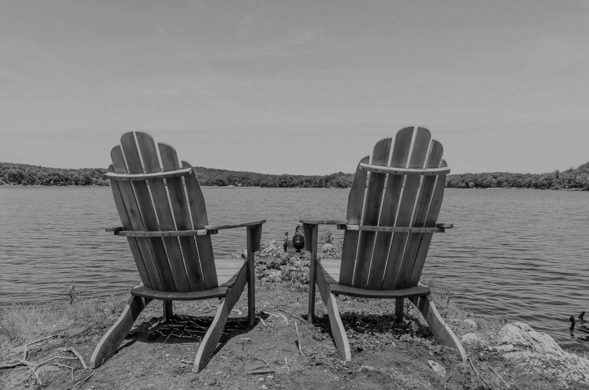 Two Adirondack chairs facing a lake, with trees along the shoreline in the background, in black and white.