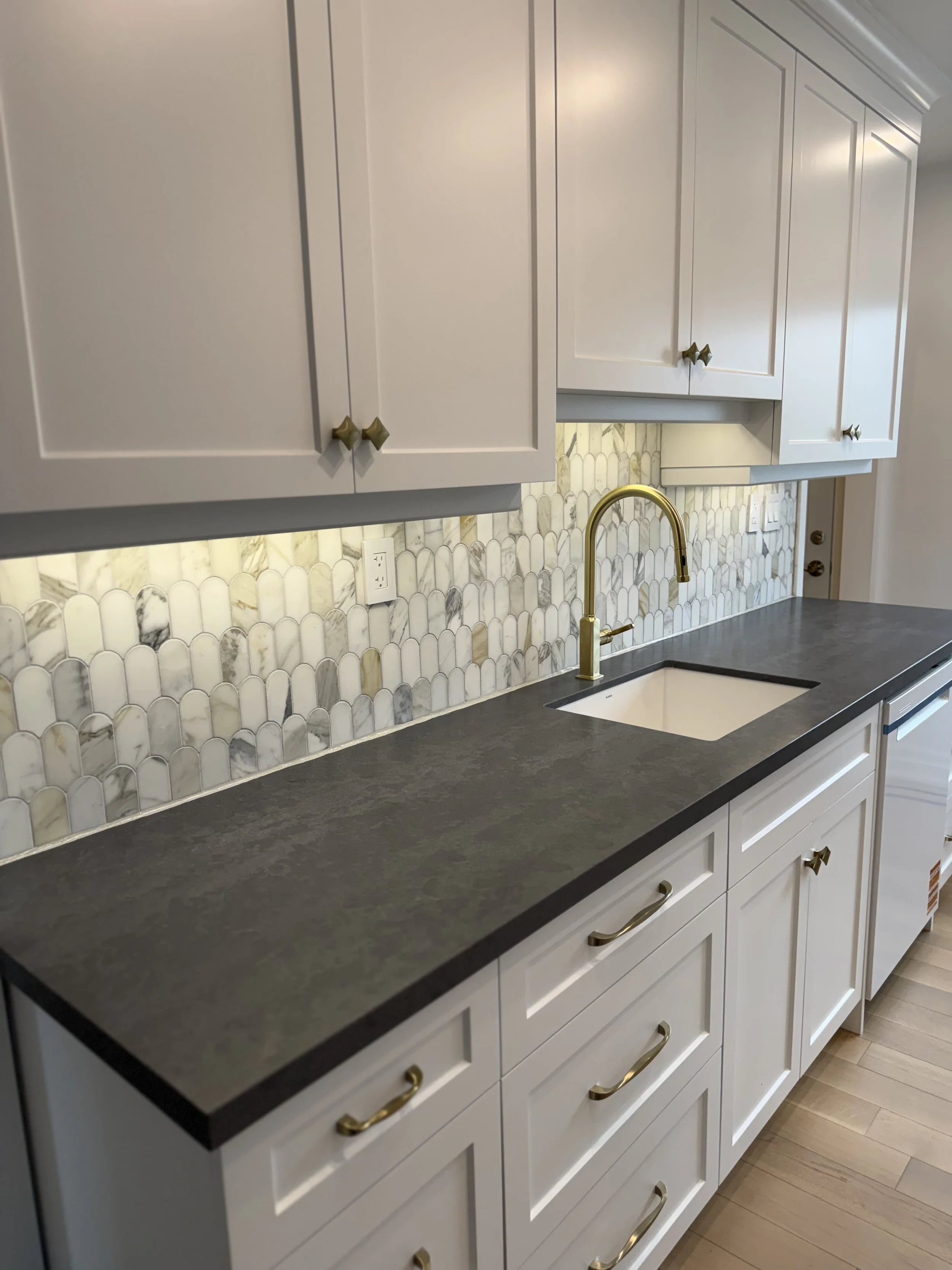 Kitchen with white cabinets, black countertop, marble backsplash, gold faucet, and wood flooring.
