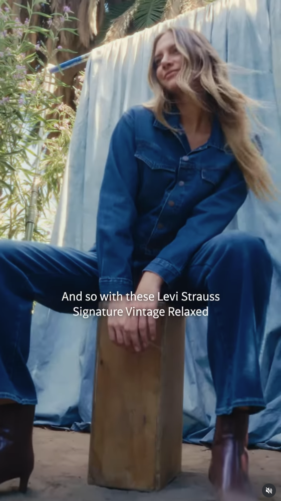 Woman sitting on a wooden box outdoors, wearing a blue denim jacket and jeans, with long hair blowing in the wind, surrounded by greenery and a blue fabric backdrop.