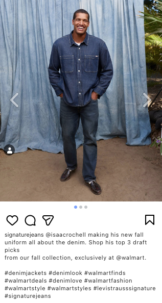 Man standing in front of a blue curtain, wearing a dark denim jacket and jeans, smiling with hands in pockets.