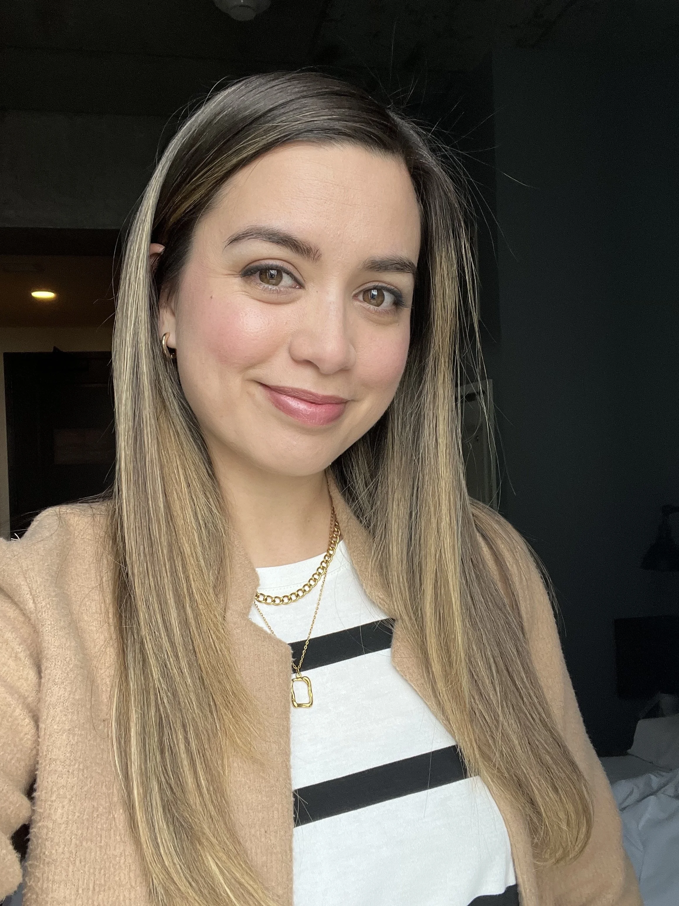 Close-up of a woman with straight, blonde hair smiling, wearing a beige jacket, layered necklaces, and a white top with black stripes.
