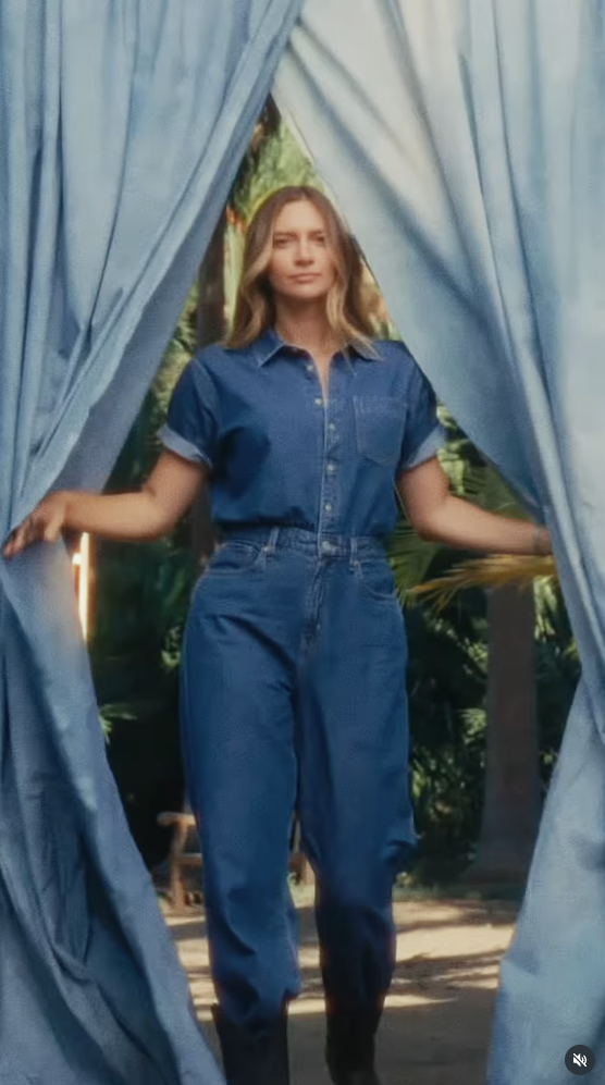 Woman in denim outfit peeking through blue curtains with greenery in the background.